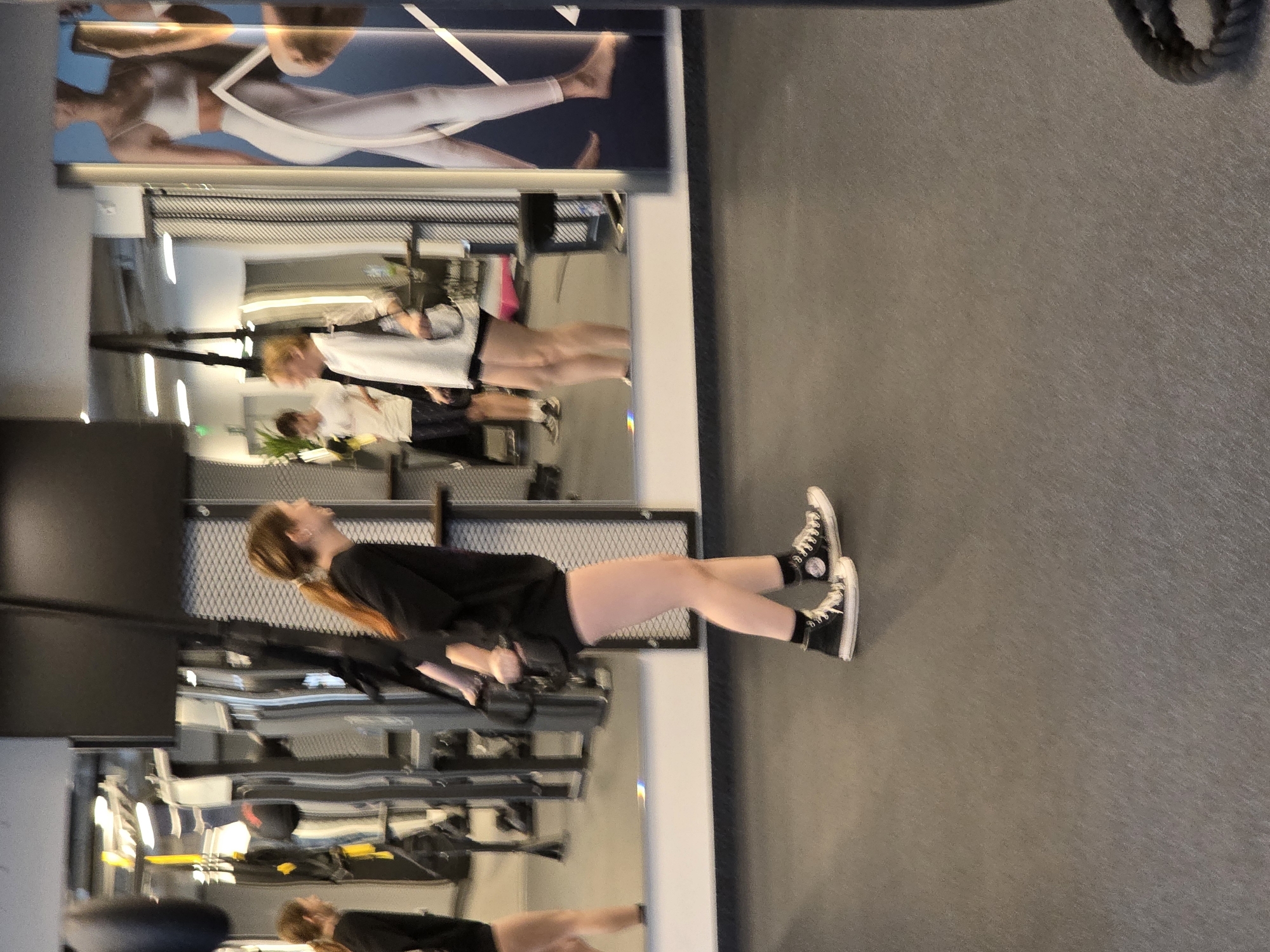 This image, taken in Plovdiv, Bulgaria, captures an indoor gym scene. The central subject is a young person, likely female, with long reddish-brown hair in a ponytail, dressed in a black short-sleeved top, black shorts, black socks, and black high-top sneakers with white trim. They are performing an exercise, leaning back at an incline while holding onto unseen equipment, possibly a suspension trainer or resistance bands. Their head is tilted upwards, and their expression appears focused.
The background is dominated by a large, wall-mounted mirror that reflects other parts of the gym. In the mirror, another person with lighter hair, wearing a white top and dark shorts, is visible further back, appearing to be engaged in an activity or standing. Additional blurred reflections of gym equipment and structures can be seen throughout the mirror.
Above the mirror, a large poster or advertisement is displayed, featuring two stylized figures, presumably female, in white activewear (sports bra and leggings), striking a dynamic pose. A white, stylized "V" shape or logo is present on the poster, but no discernible text is readable. The floor is a dark, textured gym mat, and the overall setting suggests a modern and well-lit fitness facility. The circular white logo on the side of the subject's left sneaker, characteristic of Converse Chuck Taylor All-Stars, is visible but unreadable.