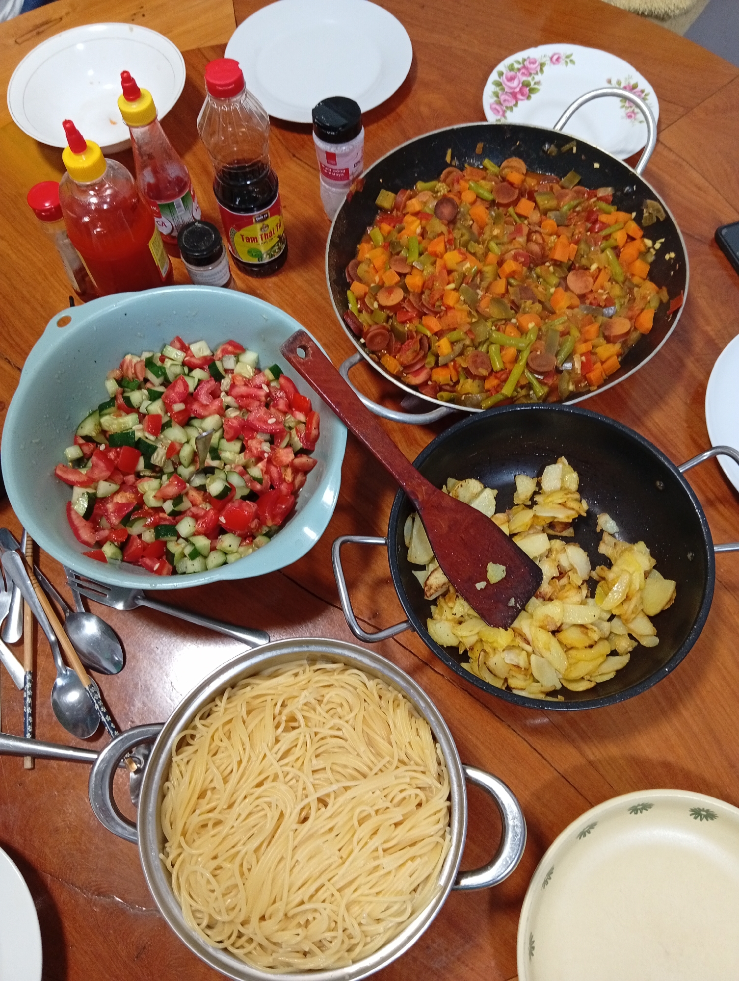 The image shows a table with a variety of dishes. A pot of cooked spaghetti sits at the bottom. Adjacent to it is a black pan containing fried potatoes with a wooden spatula. A bowl of cucumber and tomato salad is also visible. At the top, there's a pan with a mix of vegetables, possibly including carrots, green beans, and sausages. Condiments such as chili sauce and soy sauce bottles are present, along with plates of various sizes. Utensils (spoons, forks) are placed near the bowl of salad. Based on the "Tam Thai Tu" soy sauce, the image appears to have been taken in Vietnam. If I had to guess, given the fresh produce available and the style of food, it seems plausible that this image could have been taken in Da Lat, Vietnam.