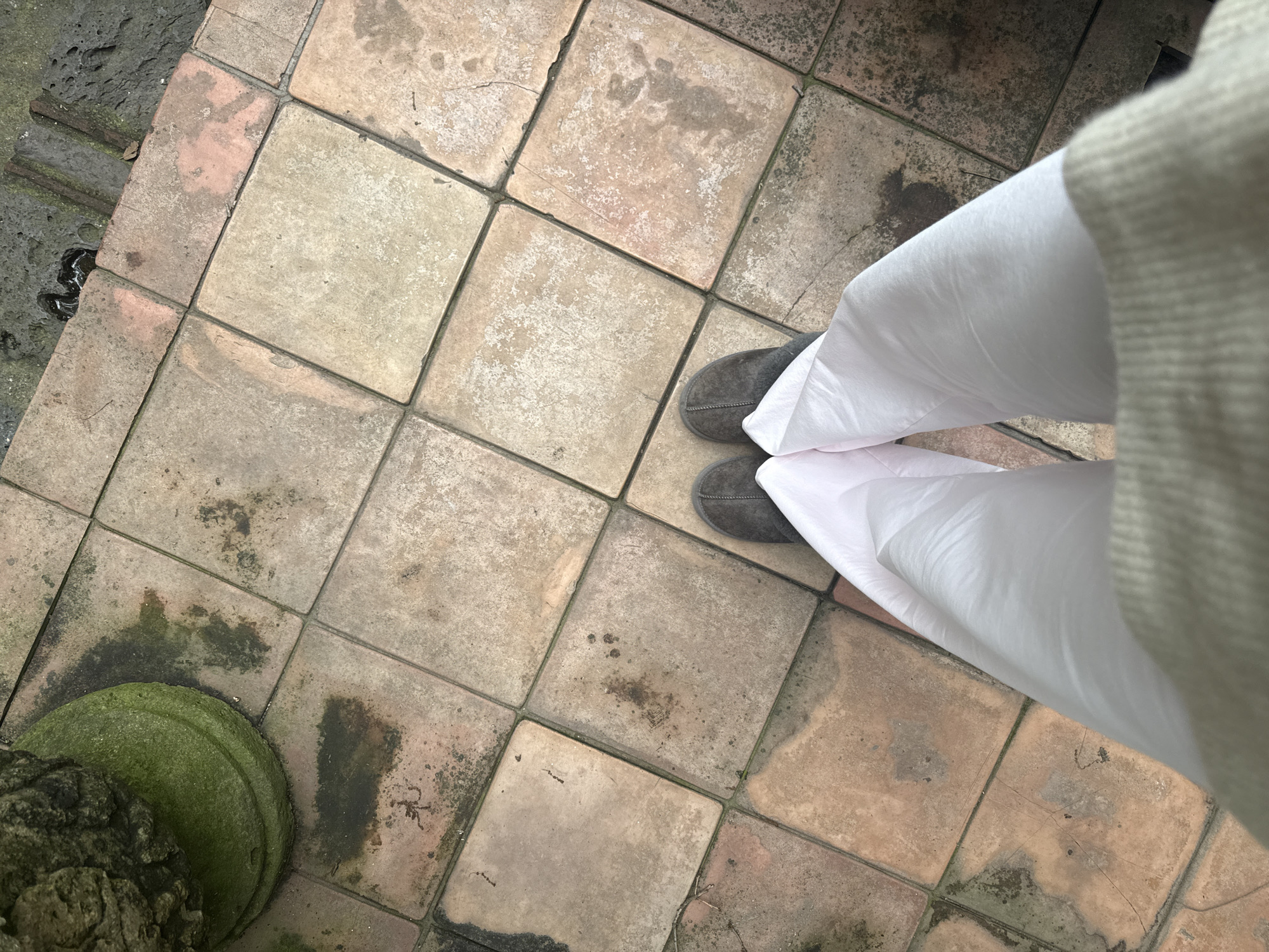 The image shows a pair of grey slippers and white pants on a tiled floor. The tiles are square and appear to be made of terracotta, with some discoloration and moss growth. A green, moss-covered stone pedestal is visible in the upper left corner. The scene is likely outdoors, possibly in a courtyard or patio.