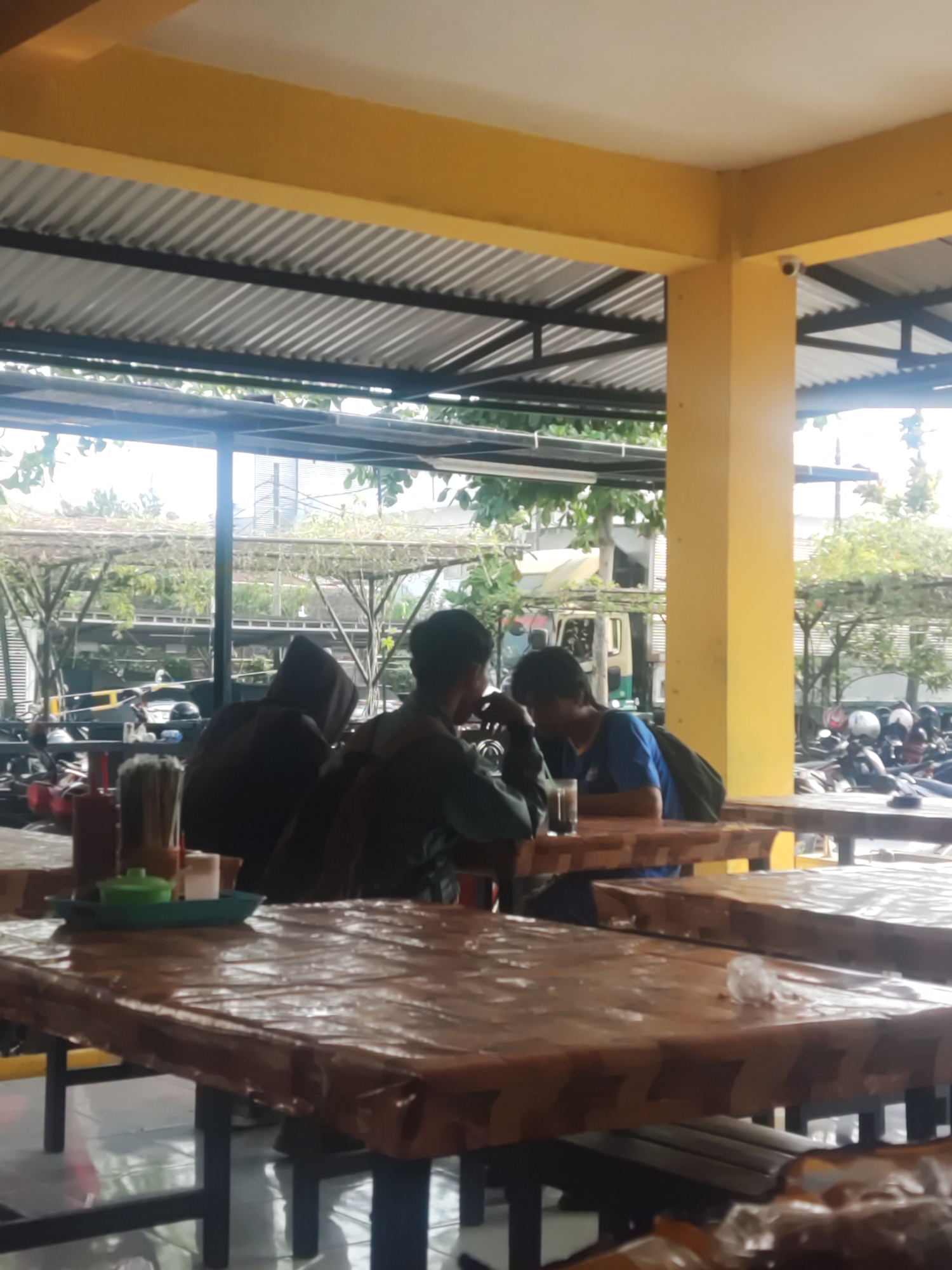 The image depicts an outdoor dining area, likely a warung or small restaurant, located in Trihanggo, Indonesia. Three individuals are seated at tables, seemingly engaged in conversation or enjoying a meal. One person, wearing a dark hooded jacket, is seated facing away from the camera. To their right, another person in a dark shirt is also facing away. A third person, wearing a blue t-shirt, is seated at a table with a glass of dark liquid in front of them. The tables are covered with brown plastic sheeting. In the background, a row of motorcycles and a yellow support pillar for the building's roof are visible. The environment suggests a casual, open-air setting.