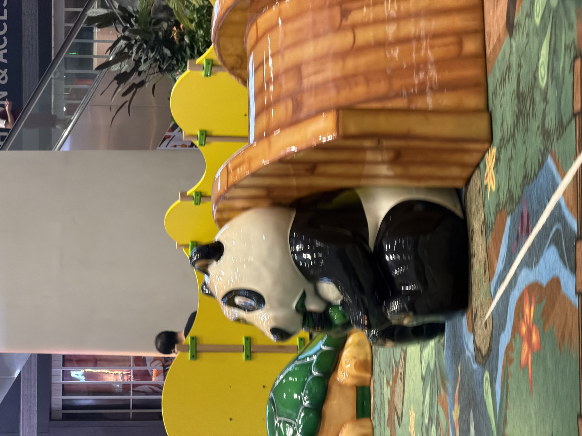 The image shows a children's play area with a panda-themed slide and a turtle-themed structure. A person's back is visible near a yellow barrier. In the background, there's an escalator and plants. The floor is covered with a colorful carpet depicting a nature scene. The overall environment appears to be indoors, possibly a mall or entertainment center.