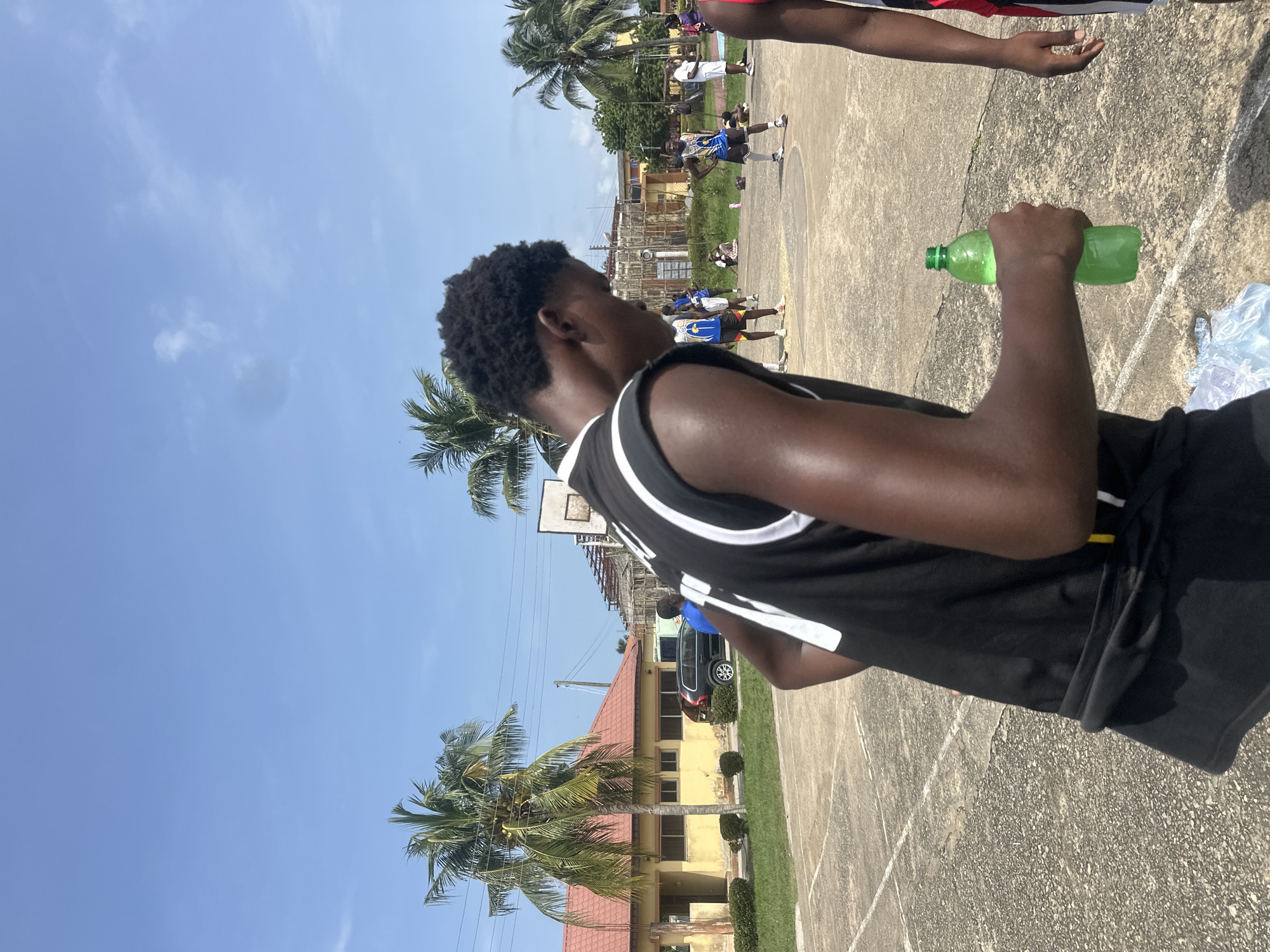 The image depicts an outdoor basketball court with several individuals present. In the foreground, a young male in a black and white basketball jersey is holding a green plastic bottle. The court surface appears to be made of concrete with white lines marking boundaries. In the background, there is a yellow building with a red roof, palm trees, and other structures, suggesting an urban or semi-urban environment. Some individuals are seen on the court, some of whom appear to be playing basketball. The sky is clear and blue, indicating a sunny day. The context provided mentions Ajara, Nigeria, which places the scene in that specific geographical location.