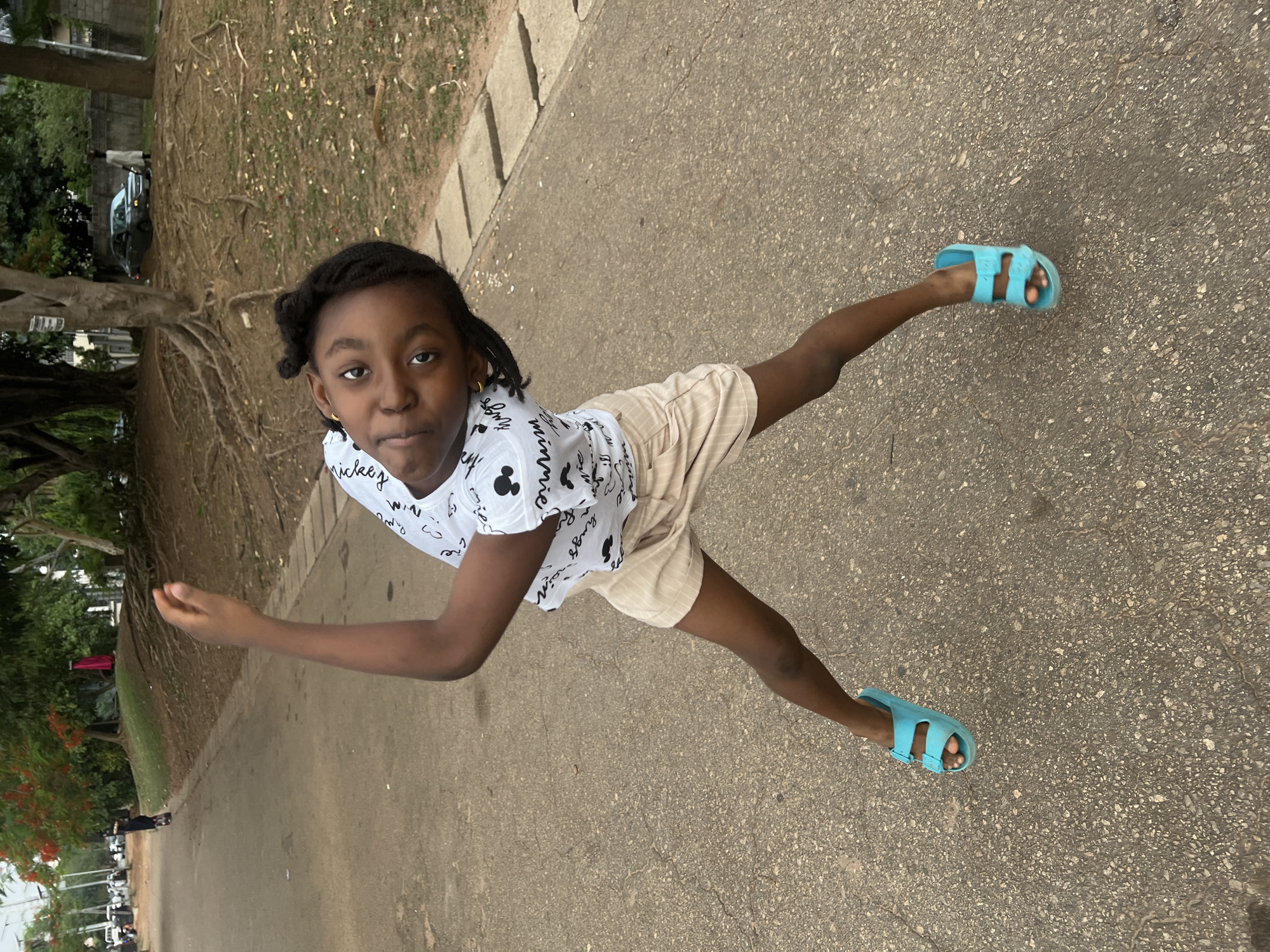 A young African girl is standing on a concrete path with grass, trees, and some parked cars in the background. She is wearing a white t-shirt with black print, beige shorts, and light blue sandals. She is holding one arm out to the side and looking directly at the camera. The location appears to be in a park or outdoor area within the city of Abuja, Nigeria, as suggested by the style of the vegetation and architecture.