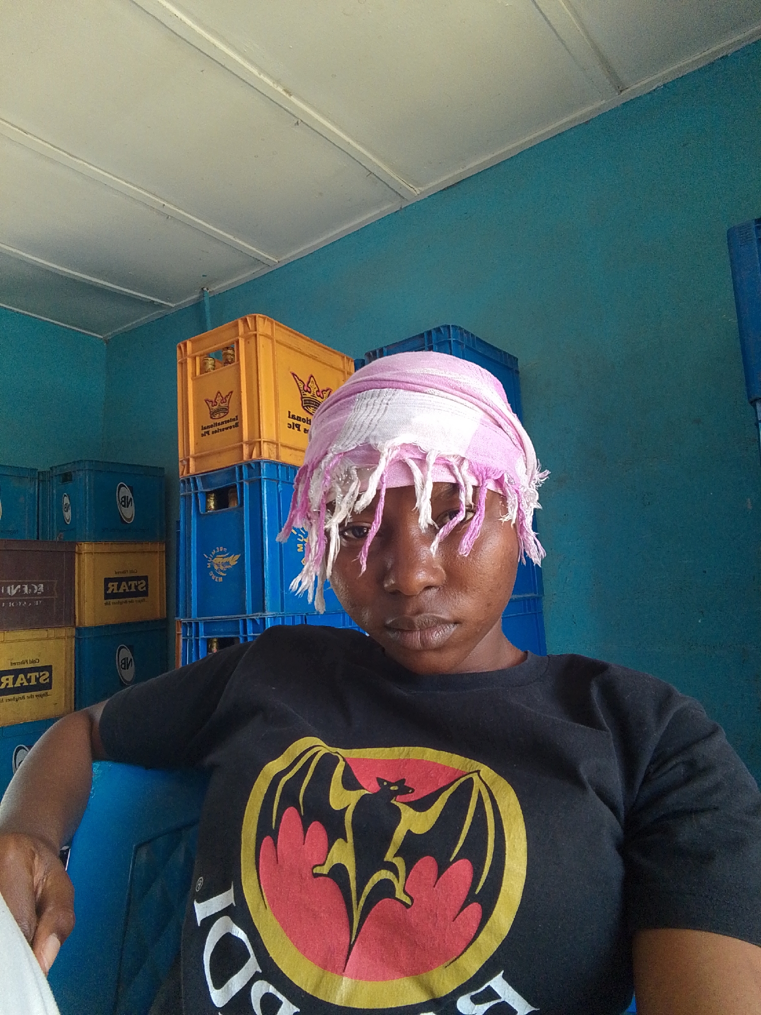 A person wearing a black T-shirt with a large Bacardi logo, and a pink and white headscarf with fringe, sits in front of stacks of yellow and blue crates, some of which appear to contain bottles. The walls are blue and the ceiling is white. Text on the crates include "STAR" and "lanol."