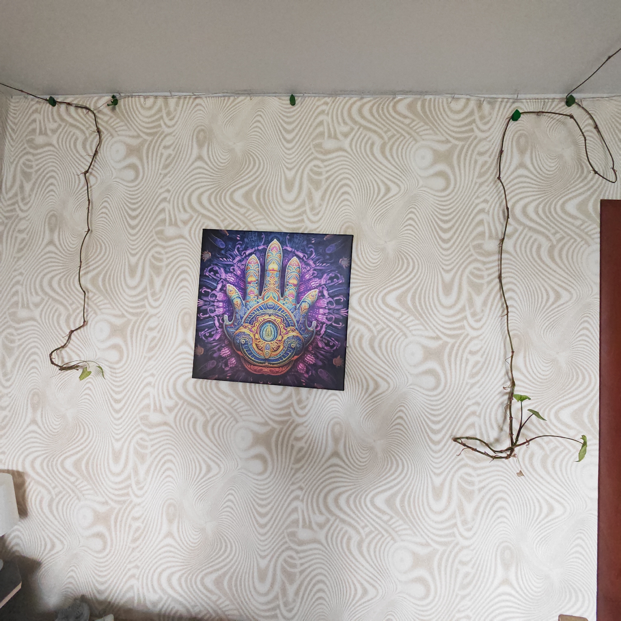The image captures an interior view of a room, showcasing a wall adorned with light-colored wallpaper featuring a distinct wavy, abstract, and almost psychedelic pattern in a slightly darker shade. Centered on the wall is a square-shaped framed artwork. This piece depicts a highly stylized and vibrantly colored hand, possibly a Hamsa or similar symbolic design, rich with intricate patterns and featuring a prominent eye-like motif in its palm. The background of the artwork is dark, primarily in shades of purple and blue with organic abstract shapes.
Two slender plant vines, adorned with small green leaves, are meticulously trained along the top edge of the wall, near the ceiling. They are held in place by small, green clips. One vine extends from the upper left corner of the image, looping downwards, while the other starts from the upper right, tracing a path downwards on the right side. A plain, light-colored ceiling is visible above the wall. To the far right, part of a brown door frame is seen. In the bottom-left corner, a light-colored, soft object is partially visible. The scene suggests a thoughtfully decorated living space in Novo-Nikol'skoe, Russia. No visible text is present in the image.
