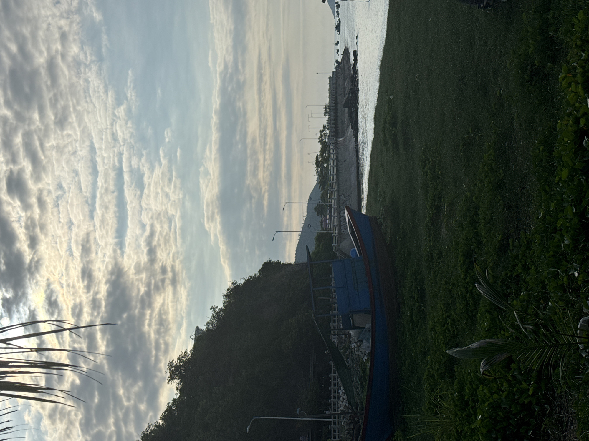 The image depicts a coastal scene in Bac Nha Trang Ward, Vietnam, under a cloudy sky with the sun peeking through. In the foreground, a weathered blue and red fishing boat rests on lush green grass. The boat has a wooden structure in the center and appears to be empty. Behind the boat, a steep, tree-covered hill dominates the left side of the frame. A concrete seawall curves along the coastline, lined with lights and a railing. Small boats are visible in the calm water beyond the seawall, and a distant hillside with structures can be seen. The overall atmosphere is tranquil and hints at early morning or late afternoon light.