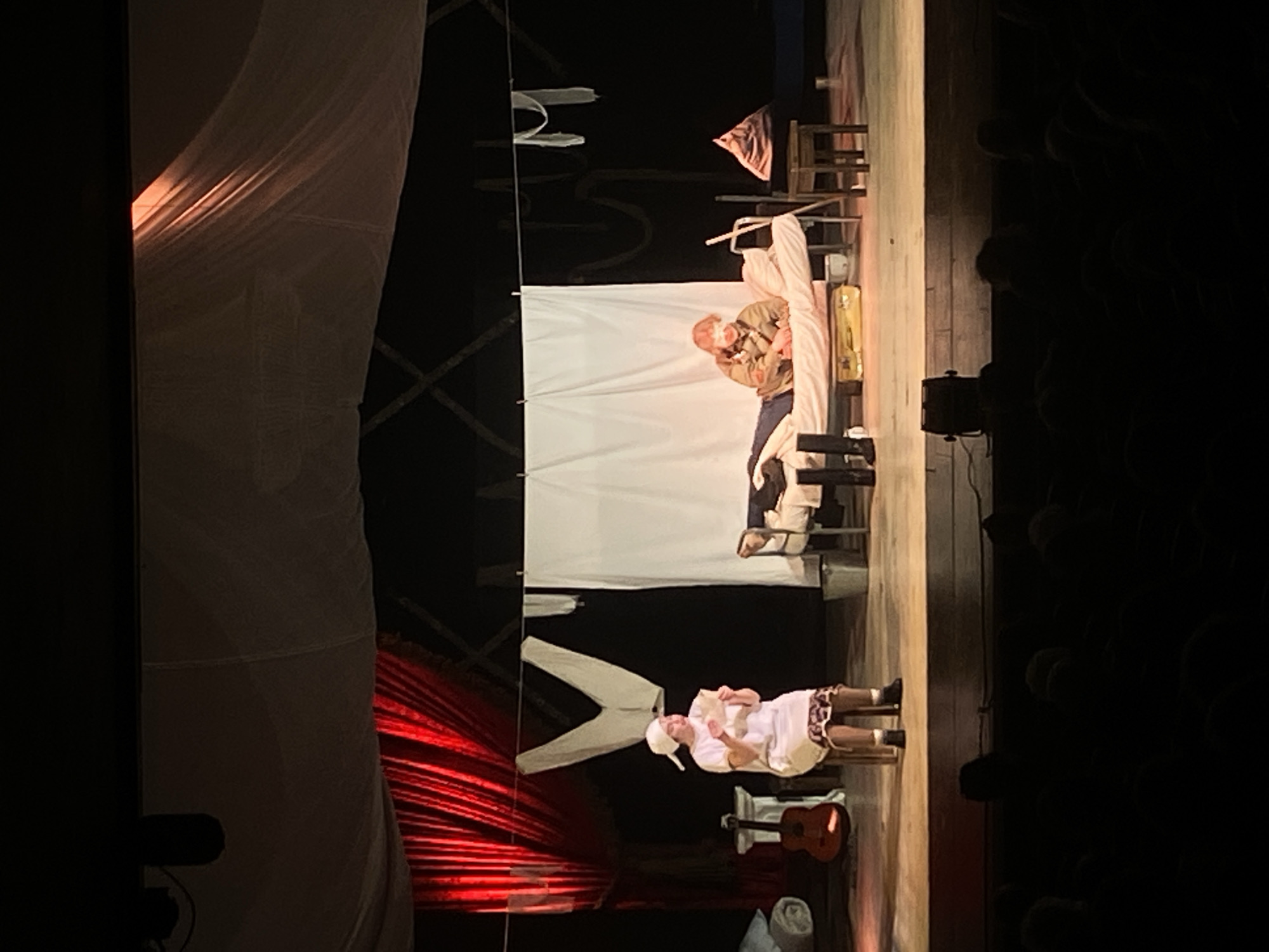 The image shows a scene from a theater play. Two actors are visible: one lying in a bed covered in white sheets and blankets, wearing a jacket and pants, and the other standing near the foot of the bed, dressed in white with a cap. A guitar rests on the floor next to a rolled-up piece of white fabric. The backdrop consists of white cloth and red curtains on the left, and on the right, part of an audience is visible. Based on visual clues, the city of Шебунино, Russia, cannot be confirmed as the location.