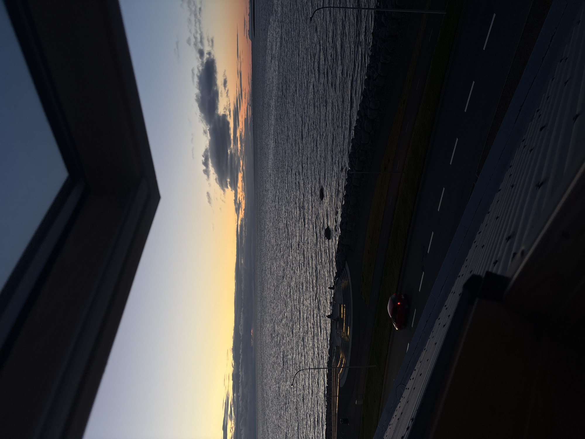 This image captures a twilight scene overlooking the ocean in Reykjavik, Iceland, likely from a window. In the foreground, a dark, corrugated roof occupies the bottom portion of the frame. Above this, a dark-framed skylight or window structure partially obscures the view of the sky and water.
The main focus of the image is the wide expanse of the ocean, with gentle ripples reflecting the soft light of the setting or rising sun. The horizon is visible, with a band of clouds tinged with orange and yellow hues from the sunset. Silhouettes of distant landmasses can be seen on the far right.
Along the shoreline, a paved walkway and a road are visible. A single red car is driving on the road. A circular, elevated structure with integrated lighting is situated between the walkway and the sea, with a few indistinct figures standing on it. A lamppost stands near the structure. The shoreline itself is lined with dark, angular rocks. The overall atmosphere is calm and serene, characteristic of a coastal evening in Reykjavik.