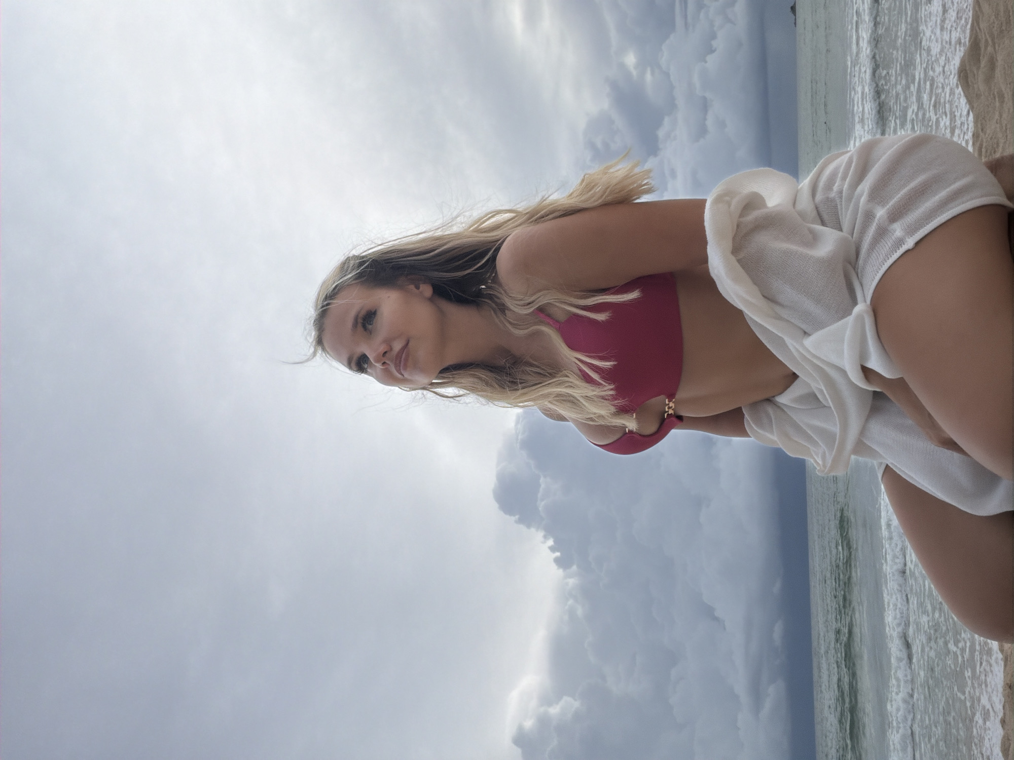 This is a vertical, medium-shot photograph taken from a low angle. The subject is a young woman with blonde hair, wearing a pink bikini top and a white cover-up. She is lying on a sandy beach with the ocean and a cloudy sky in the background. Her body is angled, and she is looking to her left.
The scene is set on a beach, likely in Choeng Thale, Thailand, given the context. The sky is filled with dramatic, heavy clouds, suggesting overcast weather, possibly before or after a storm. The ocean waves are visible, and the sand is light in color.
The woman appears relaxed, with her gaze directed away from the camera. Her expression is serene. The time of day is unclear due to the overcast conditions, but it appears to be daytime.
There is no visible text in the image.