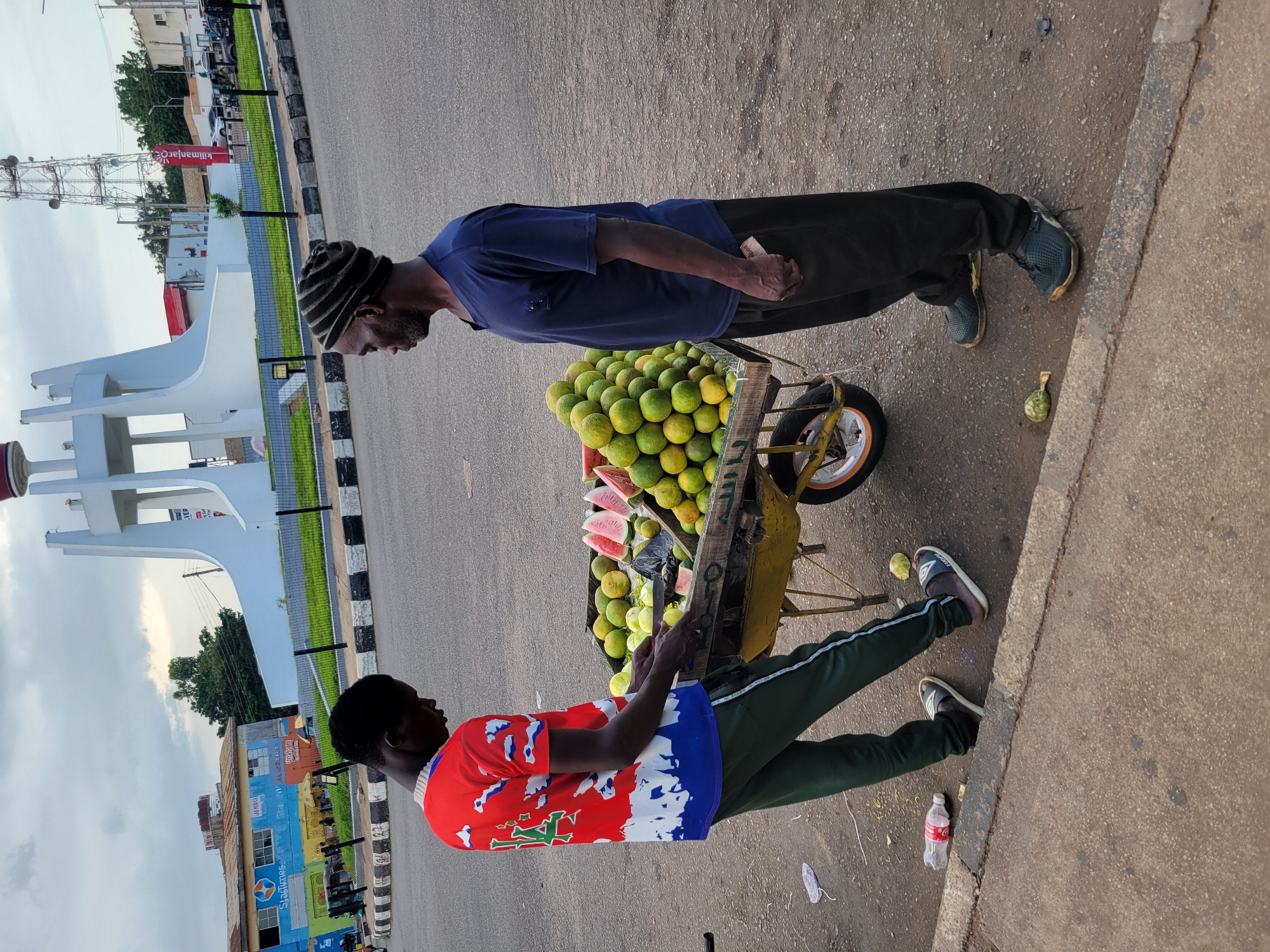 In Barakin Lerawi, Nigeria, two men stand next to a fruit cart laden with oranges and sliced watermelons. The cart, a yellow wheelbarrow, is positioned on the asphalt road. The younger man, wearing a red shirt with a white and blue abstract design and dark green track pants, appears to be interacting with the fruit. The older man, dressed in a navy blue t-shirt and dark pants, and a striped knit cap, stands to the right. In the background, a white monument with a red top is visible, along with a cellular tower. Buildings, including one with "StarTimes" signage, and parked vehicles contribute to the urban landscape. The sky is overcast with clouds. Litter, including a plastic bottle, is scattered on the ground.