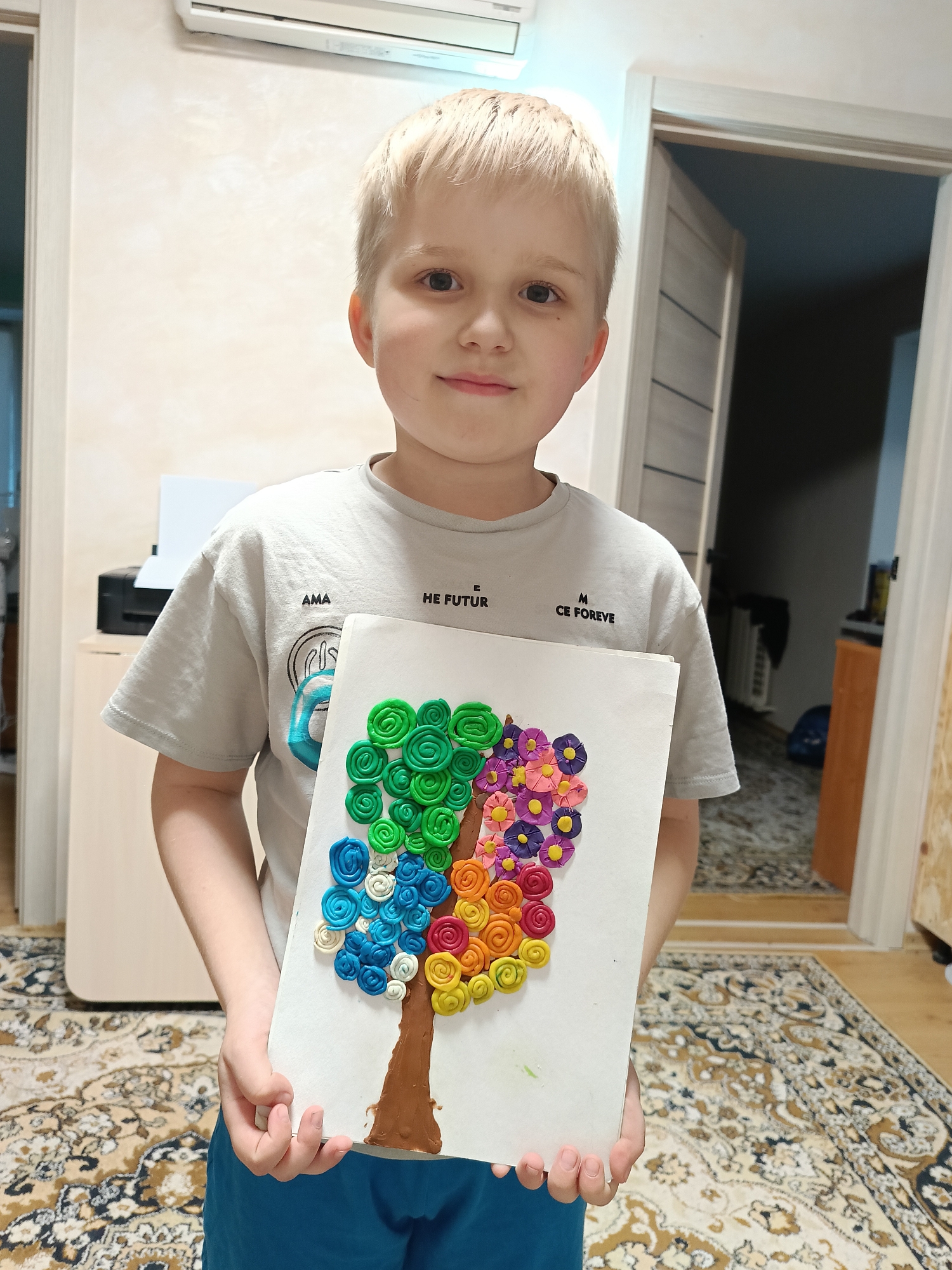 A fair-skinned boy with short blonde hair is holding up a piece of artwork he has made. The artwork depicts a tree with a brown trunk and a canopy made of rolled clay in various colors, arranged in vertical bands of green, blue, yellow, orange, pink, and purple. The boy is wearing a grey t-shirt with text on it, and blue shorts are visible.
The background is a domestic interior. Behind the boy, there is a wall painted in a light neutral color, with an air conditioner mounted high up. To the left, a printer sits on top of a light-colored cabinet. An open doorway to the right reveals a glimpse into another room. The floor is covered in a patterned rug with brown and beige tones.
While the city of Chaikovsky, Russia, cannot be definitively identified from the image alone, the domestic interior setting and general appearance of the objects and individuals present are consistent with many residential settings in Russia.