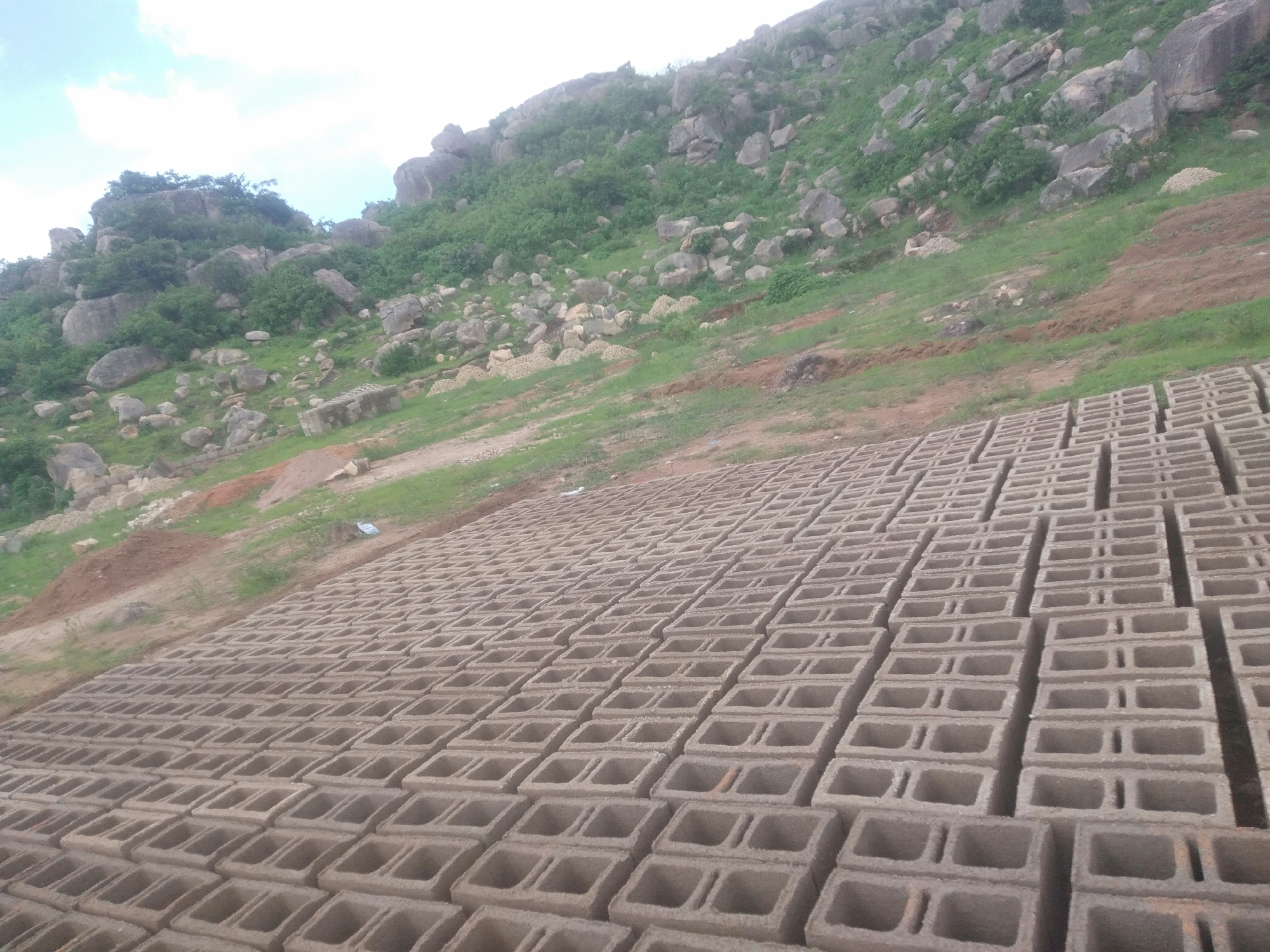 The image depicts a construction site with a large quantity of concrete blocks neatly arranged in rows on a sloped, grassy area. In the background, a rocky, tree-covered hillside rises under a bright, partly cloudy sky, suggesting an outdoor daytime setting. The foreground is dominated by the orderly stacks of building materials. The blocks are uniform in size and shape, with a hollow, rectangular design, indicating they are likely intended for construction. The ground beneath the blocks appears to be packed earth, with patches of green grass and some scattered smaller rocks. The overall scene conveys a sense of ongoing construction or preparation for a building project in a natural, somewhat rugged environment. There are no people visible in the frame. The lighting suggests it is daytime, possibly late morning or early afternoon, given the brightness of the sky and the shadows cast by the blocks. The location appears to be rural or semi-rural, given the natural landscape and the absence of urban structures.