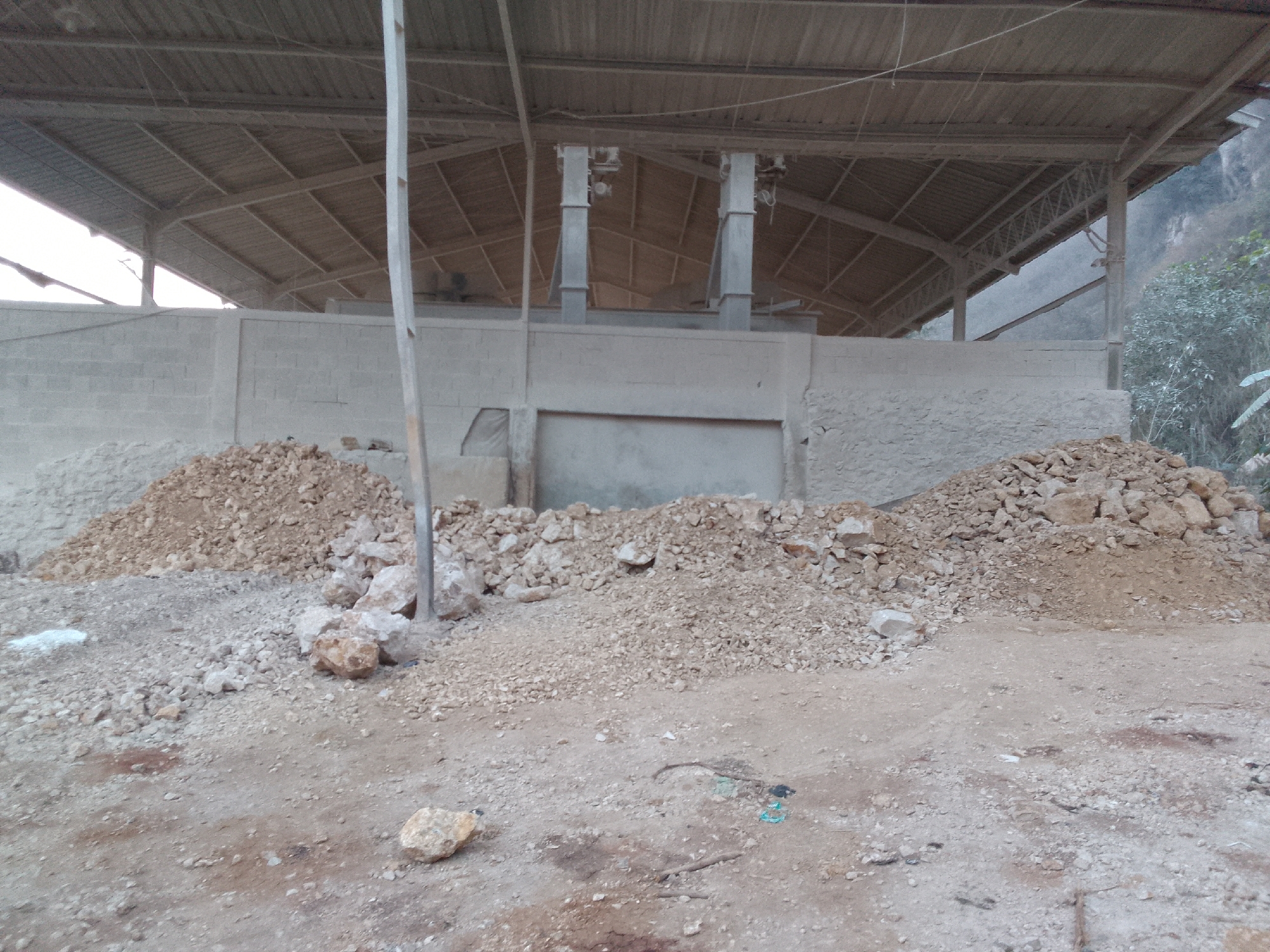 This is a full, eye-level, outdoor shot of an industrial area or quarry in Gunungmasigit, Indonesia. The foreground is a dirt path with scattered rocks and some debris, including a blue string. There are three large piles of rocks and dirt on the left and right sides of the image, with a smaller pile in the middle. In the background, there is a long, open-air building with a corrugated metal roof supported by a wooden structure. A wall made of concrete blocks runs along the front of the building. Two large, cylindrical metal structures, possibly part of a conveyor system, are visible within the building. The area appears dusty and somewhat unkempt, suggesting an active or recently used industrial site. The natural environment is visible on the far right with a steep, rocky hillside covered in vegetation. The sky is overcast and bright. There are no people visible in the image.
