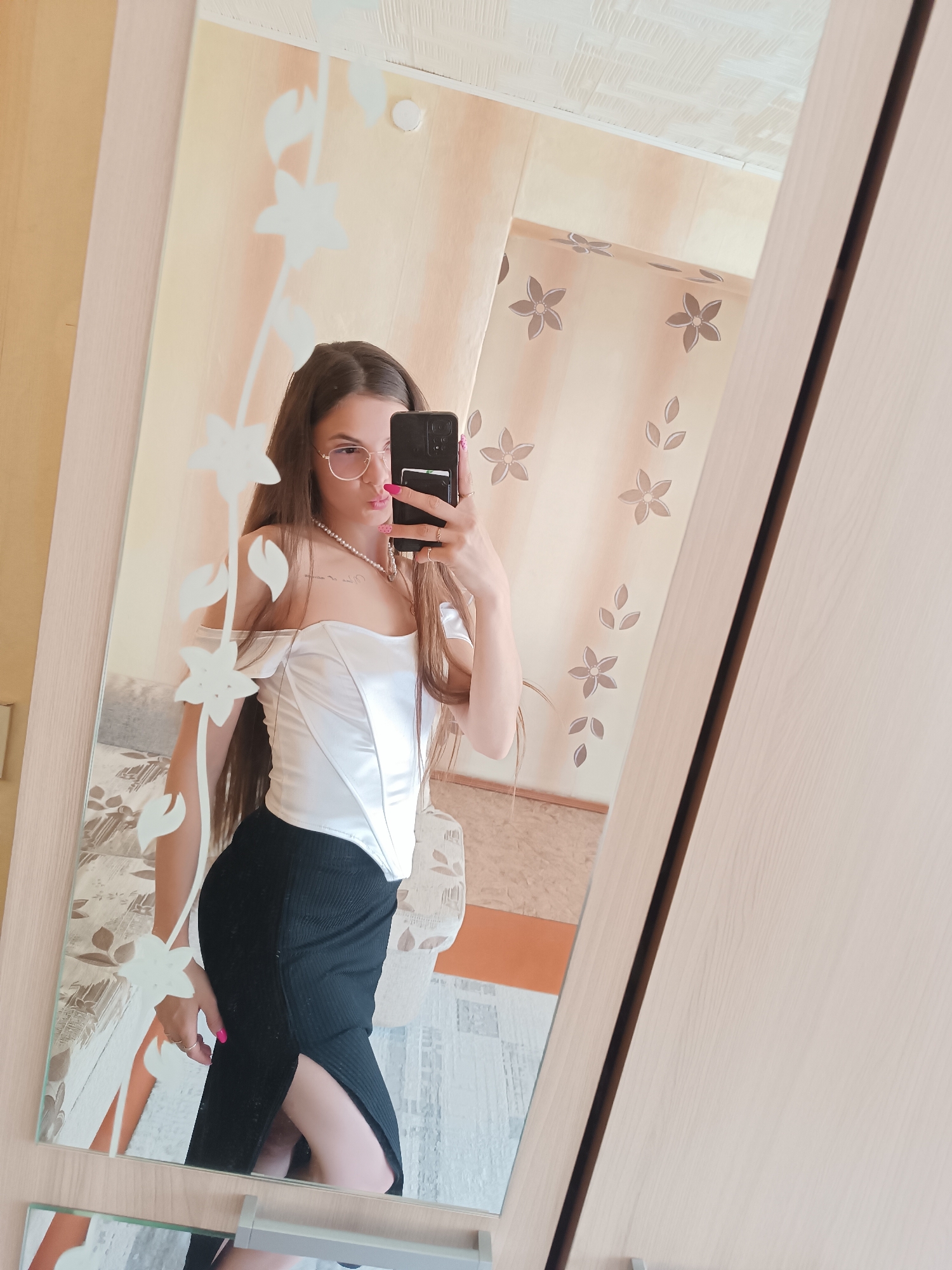 A young woman stands in front of a mirror, taking a selfie with her smartphone. She is wearing round-framed glasses, a white off-the-shoulder top, and a black skirt with a slit. She also wears a pearl necklace. The mirror is decorated with a floral design. The background shows a room with floral-patterned wallpaper and furniture, presumably in a residential setting. The location is Unknown, Unknown.