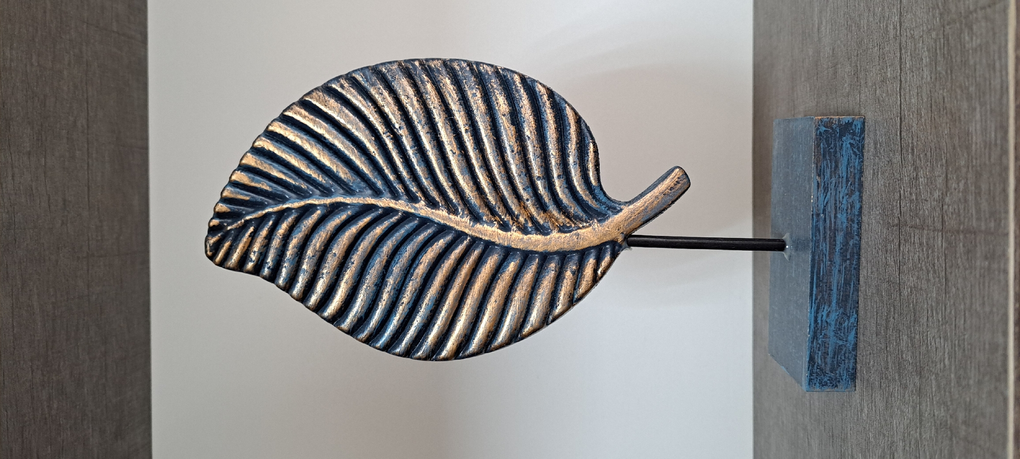 A dark blue and gold leaf sculpture is displayed on a dark gray surface in front of a white wall. The sculpture is a stylized representation of a leaf with prominent veins, rendered in textured dark blue with accents of metallic gold. It is mounted on a thin black metal rod, which is attached to a dark blue rectangular base. The base rests on a textured surface that appears to be a wooden shelf. The background wall is plain white, and above the white wall, a dark horizontal surface, possibly a shelf or cabinet, is visible. The lighting suggests a studio or indoor display environment, with no visible signs of the city of Yessentuki, Russia.