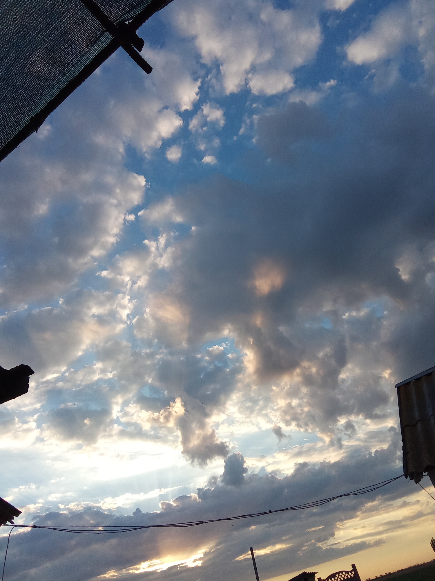 This is a photograph of a cloudy sky over Oleksandrivka, Ukraine. The image is taken from a low angle, looking upwards. In the foreground, a dark, textured surface, possibly a shade cloth or tarp, occupies the upper left corner. To the right and bottom right, parts of roofs are visible, one appearing to be made of corrugated metal. The sky is filled with a mix of white and dark grey clouds, with patches of blue sky visible between them. Sunlight is breaking through some of the clouds, creating bright rays and illuminating sections of the cloud cover with warm, golden hues. A dark wire stretches horizontally across the lower portion of the image. The overall impression is of a dynamic sky during sunset or sunrise.
