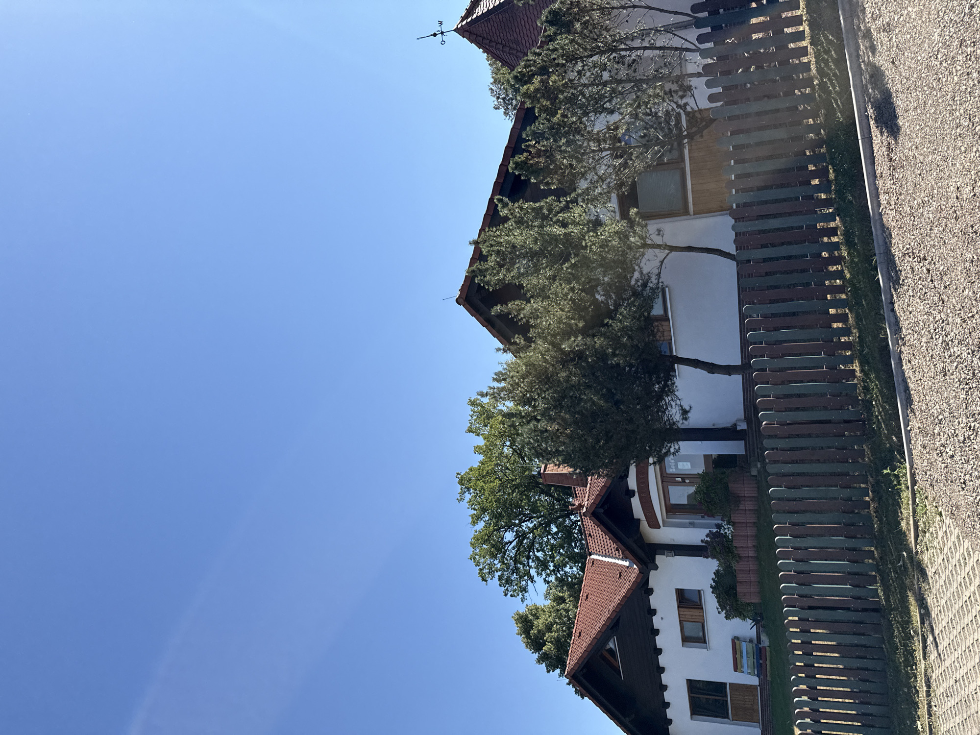 The image shows a white, two-story building with a red-tiled roof, situated in Bad Sachsa, Germany. The building has dark trim around the roofline and windows. A colorful wooden bench is visible on the left side of the building, and a wooden fence runs along the front of the property. The fence is made of vertical wooden slats painted in alternating shades of green and brown. In front of the fence, there is a patch of grass, and beyond that, a gravelly area and a paved walkway. Several trees, including conifers and deciduous trees, are planted around the building and in front of it. The sky is clear and blue, indicating a sunny day. A weathervane is visible on the roof of the building on the right. There are no people visible in the image.