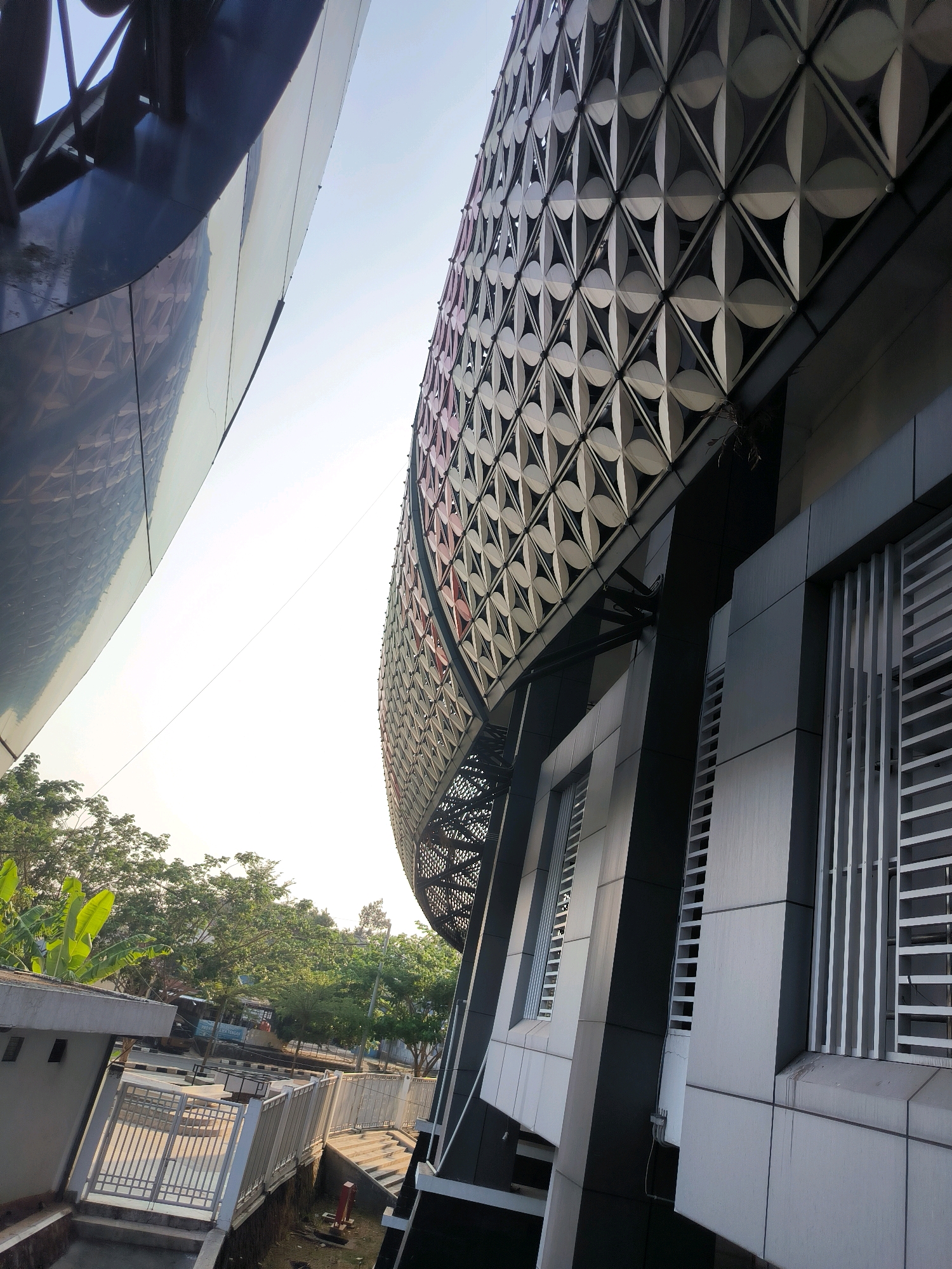 This is an outdoor, ground-level, medium-shot, high-angle, daylight photograph of a modern building with an intricate facade. The building appears to be constructed of dark, reflective panels and a patterned metallic screen. To the left, another building or structure with a curved, dark, reflective surface is visible. The patterned screen on the main building is composed of repeating geometric shapes resembling overlapping circles, creating a visually striking texture. The building also features vertical slats on some sections, possibly for ventilation or aesthetic purposes. In the background, lush green trees and some vegetation are visible, suggesting an urban or suburban setting. The sky is clear and bright, indicating a sunny day. The scene captures a portion of a building's exterior, focusing on its architectural details and the surrounding environment. The image is likely taken in Semarang, Indonesia, given the architectural style and the presence of tropical vegetation.