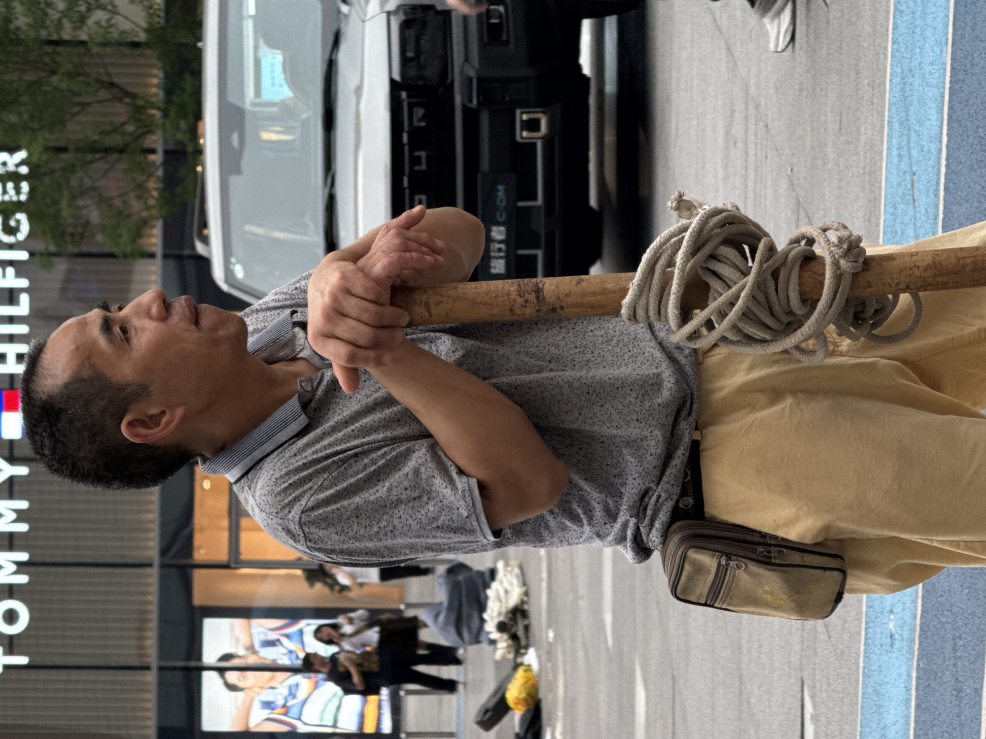 An Asian man rests a bamboo pole holding a rope on his shoulders. He wears a grey patterned shirt and tan pants, with a small bag attached to his belt. The scene is outdoors in Nan'an District, China, evidenced by the Mandarin writing on the vehicle in the background. The man is positioned near a Tommy Hilfiger store. Other pedestrians are visible in the background. A white vehicle is parked nearby. The ground is paved.