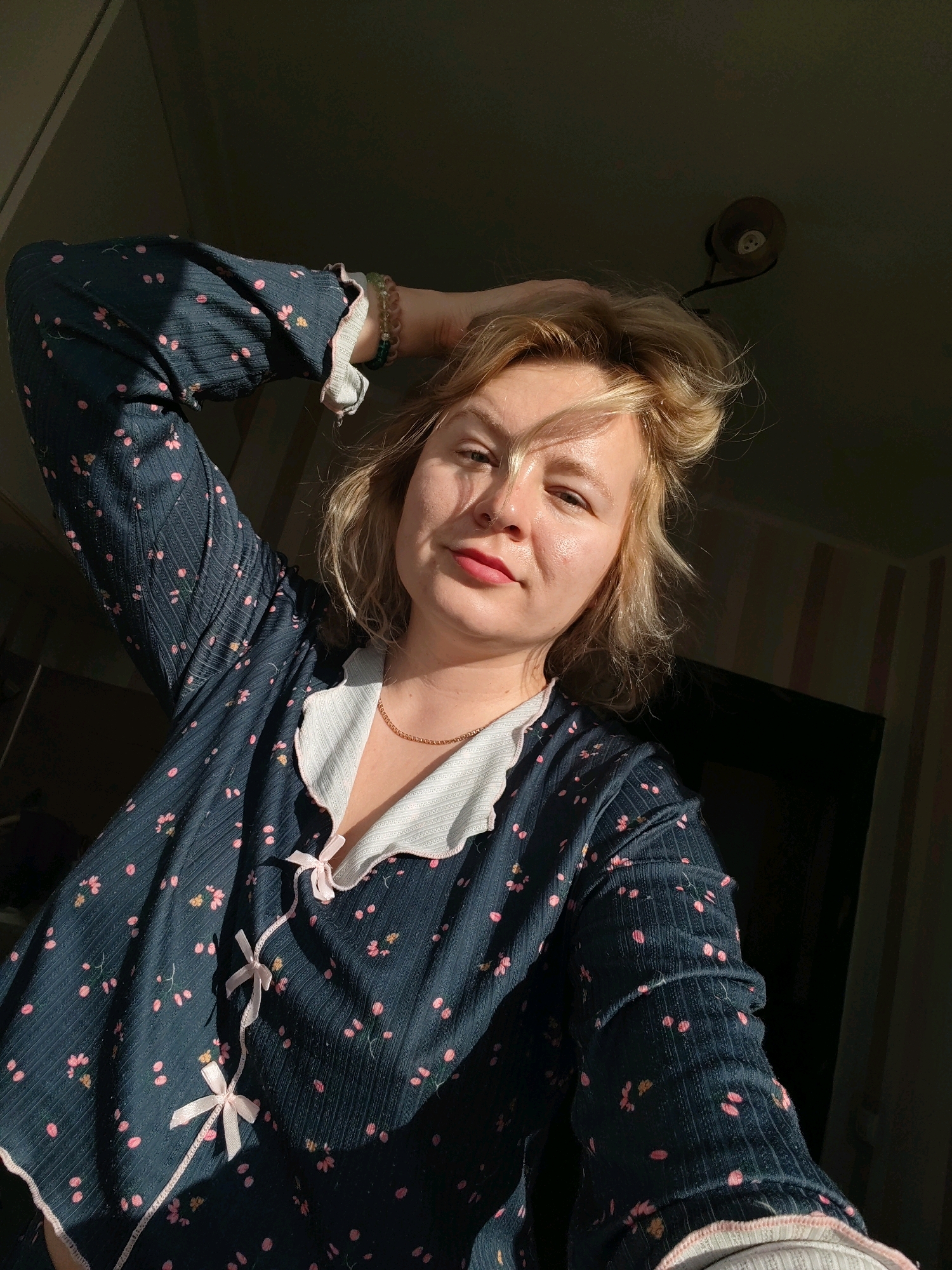 A young Caucasian woman with blonde hair pulled back is taking a selfie in what appears to be a dimly lit room. She is wearing a dark blue pajama top with a pink floral pattern and a white collar. The woman is looking at the camera and has her left arm raised, with her hand resting on her head. Sunlight is casting shadows across her face and the room. The background is out of focus, but hints of striped wallpaper and a ceiling light fixture are visible. The image provides no specific details about the location in Izhevsk, Russia.
