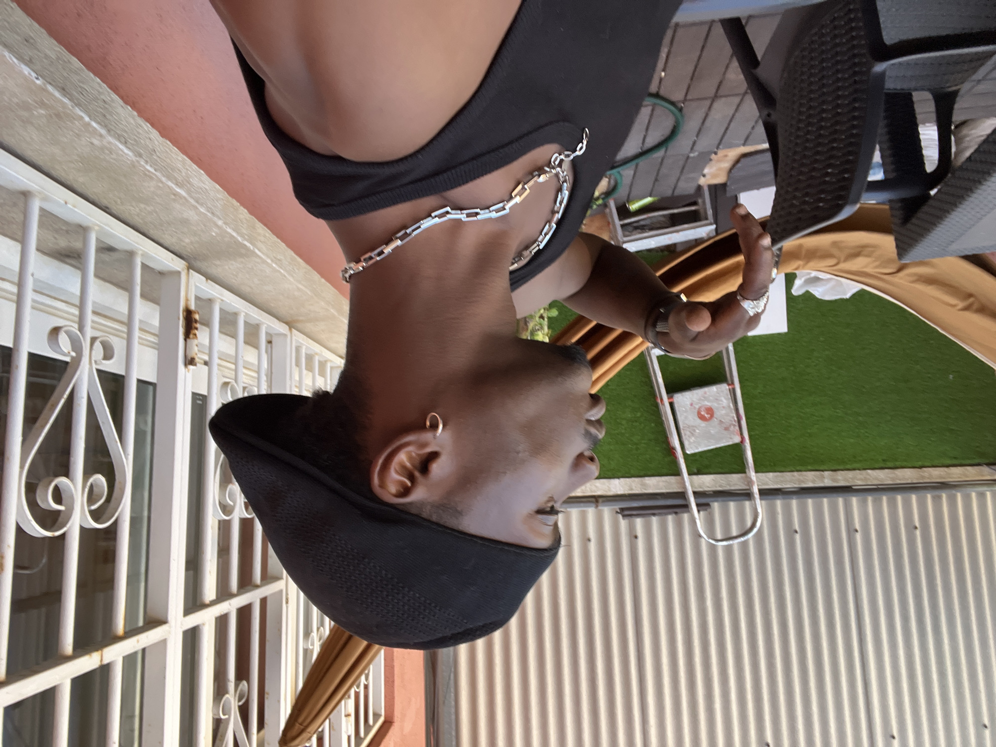 A person, an adult male with dark skin, is the central subject, captured in a side profile from the chest up. He wears a black sleeveless top and a prominent silver-toned chain necklace with rectangular links. A black, textured cap or beret covers his head, and a small gold hoop earring is visible in his left ear. On his raised right hand, he wears a silver-toned ring on his index finger and a thin, dark bracelet on his wrist. His expression is neutral as he looks to his right (viewer's left).
The setting is an outdoor patio or balcony, well-lit suggesting daytime. To the left, white metal security bars with decorative scrollwork are seen against a reddish-brown wall and a lighter-colored overhead structure. To the right, various outdoor elements include a dark, textured chair, green artificial turf, a silver ladder, corrugated metal siding, and a large tan umbrella or canopy. A green garden hose is also partially visible in the background. The scene suggests a relaxed outdoor environment, consistent with an urban residence or establishment in Barcelona, Spain. There is no specific activity depicted other than the person posing for the photograph. No visible text is present in the image.