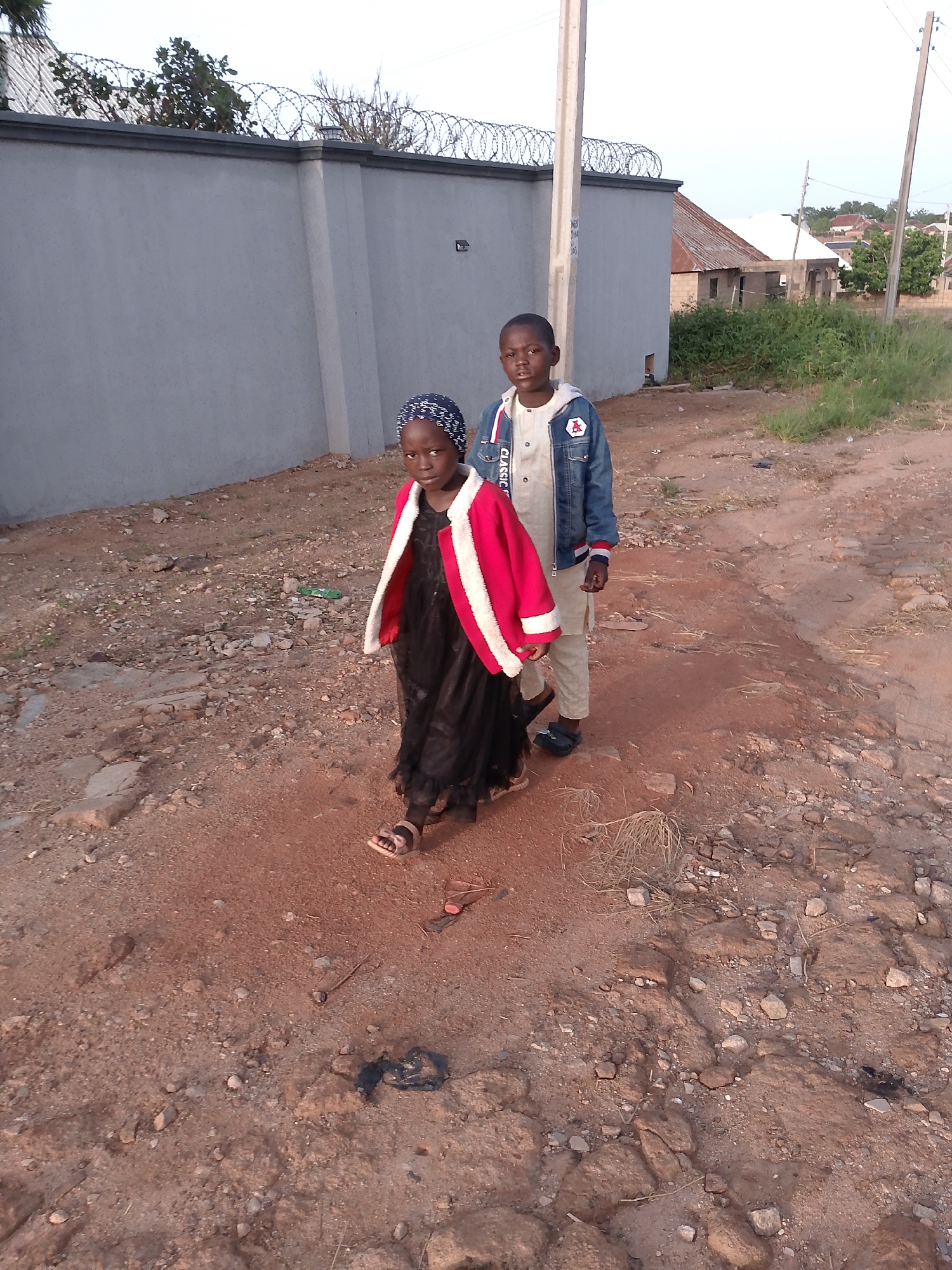 In Barikin Gidar Kanat, Nigeria, two children are walking along a dirt road beside a gray concrete wall topped with barbed wire. The child on the left, a girl wearing a black dress and a red and white jacket with a headscarf, walks ahead and to the left. The child on the right, a boy wearing a light-colored outfit and a blue denim jacket, follows behind and to the right. The ground is a mix of reddish-brown dirt and scattered stones. In the background, there are some residential buildings, a green grassy area, and vegetation. The sky is overcast.