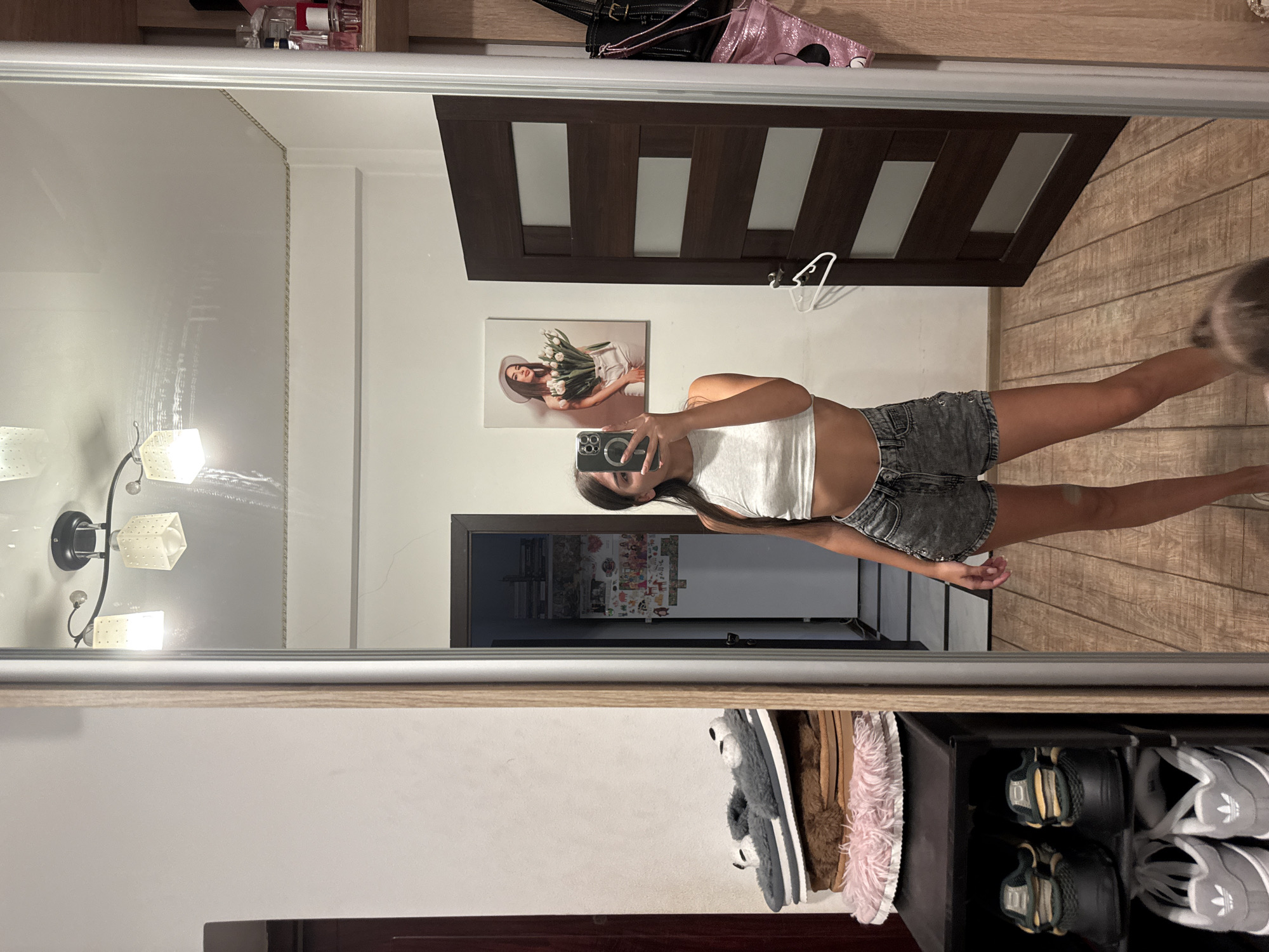 A young woman stands in front of a full-length mirror, taking a selfie. She is wearing a white crop top and denim shorts. Her reflection shows her in a room with wooden floors and a dark wooden door. To the left of the mirror, there is a shelving unit with several pairs of shoes. Above the door, a framed picture of a woman holding tulips hangs on the wall. The room appears to be a bedroom or dressing area.
