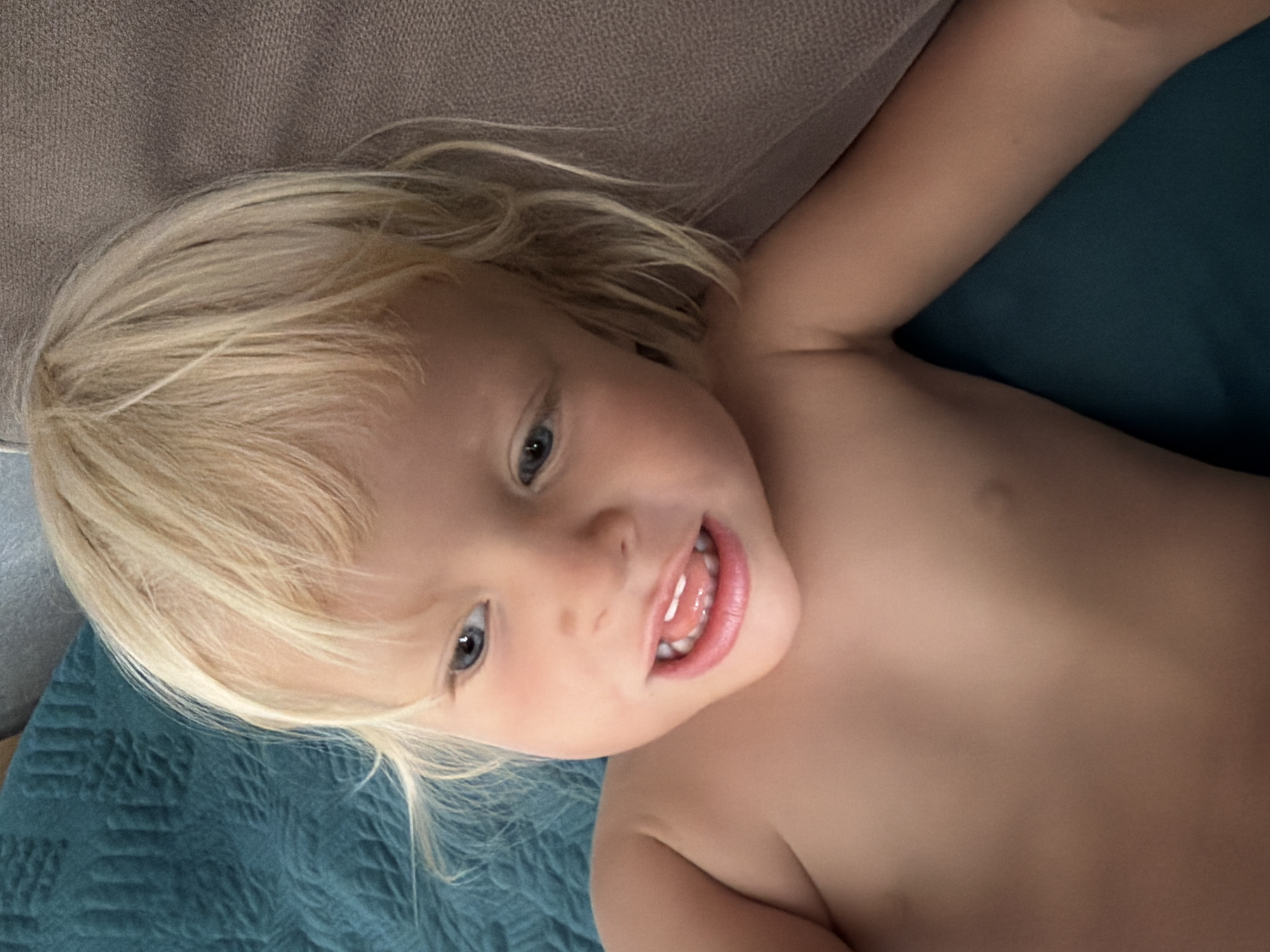 A close-up, eye-level shot shows a Caucasian toddler with blond hair and blue eyes. The toddler is lying down and looking to the left of the frame with an open mouth, revealing teeth and tongue. The child's bare shoulders and chest are visible. The background consists of a teal-colored quilted blanket and a brown textured surface, possibly a couch. The lighting is bright, casting a warm glow on the child's skin. The image appears to have been taken indoors, in a residential setting. There are no identifying landmarks or indications of the specific location in Sochi, Russia.