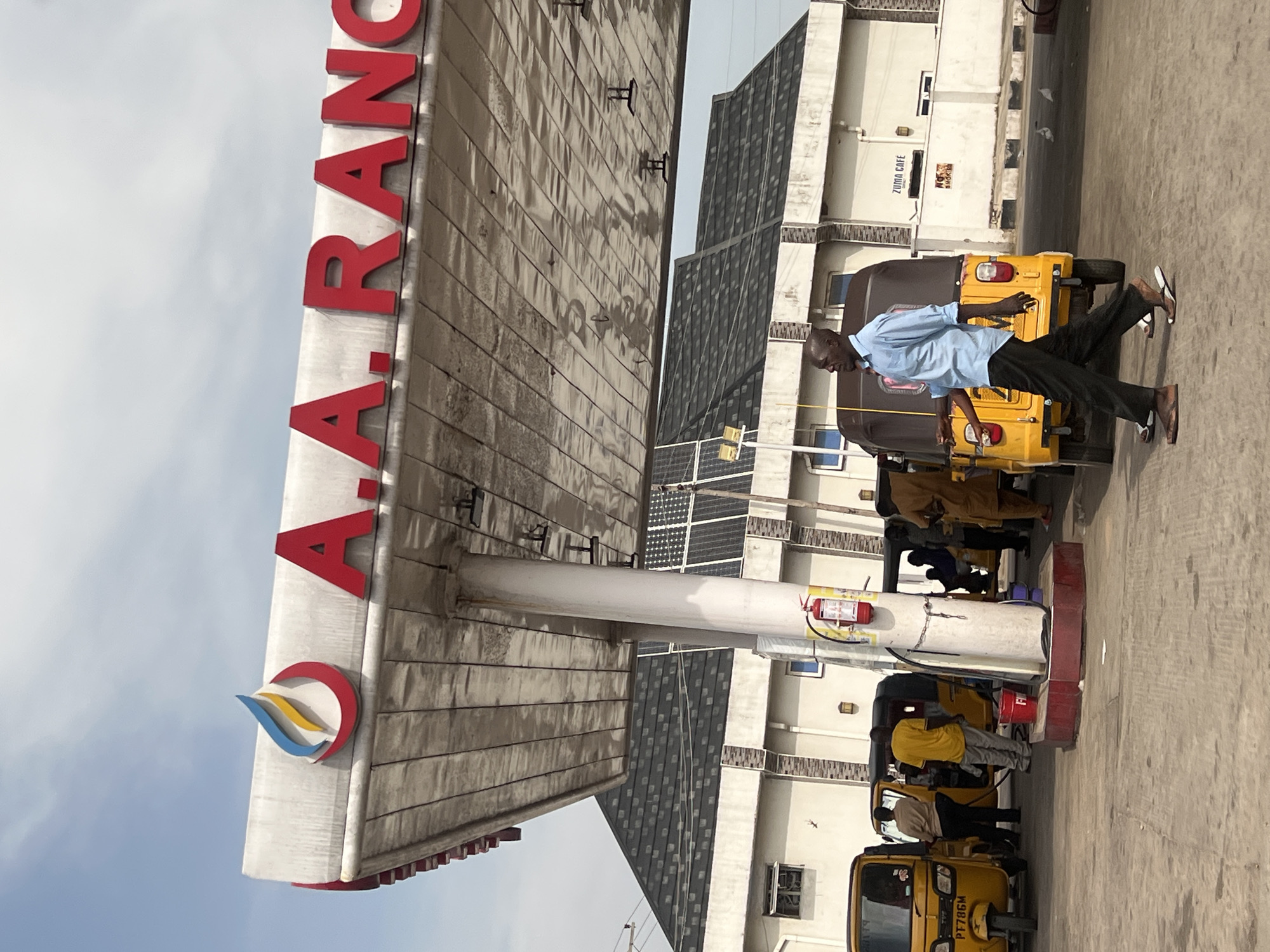 The image depicts an outdoor scene at what appears to be a gas station in Jalingo, Nigeria. A prominent red and white sign with large red lettering, partially visible, reads "A.A. RANO". Below the sign, a person in a light blue shirt and dark pants is standing next to a yellow auto-rickshaw, possibly a tuk-tuk. The auto-rickshaw has "PT786M" visible on its license plate. In the background, there are buildings with multiple stories, some featuring solar panels on their roofs. Another auto-rickshaw, also yellow, is parked nearby, with people gathered around it, suggesting refueling or interaction. A red fire extinguisher is mounted on a pillar. The ground is paved. The lighting suggests it is daytime, and the sky is overcast. The overall impression is of a functional, everyday scene at a service station.