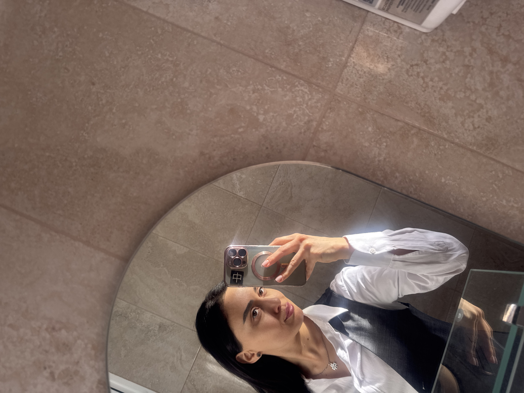 A person with dark hair is taking a selfie in a mirror. The person is wearing a white button-down shirt and a dark vest. The phone is held up to their face, obscuring some of their features. The background is a tiled wall with a warm, neutral tone. The scene is indoors, likely in a bathroom or similar space, but the exact location in Tbilisi, Georgia cannot be determined from the visual information alone. There is a white rectangular object on the wall, above the mirror.