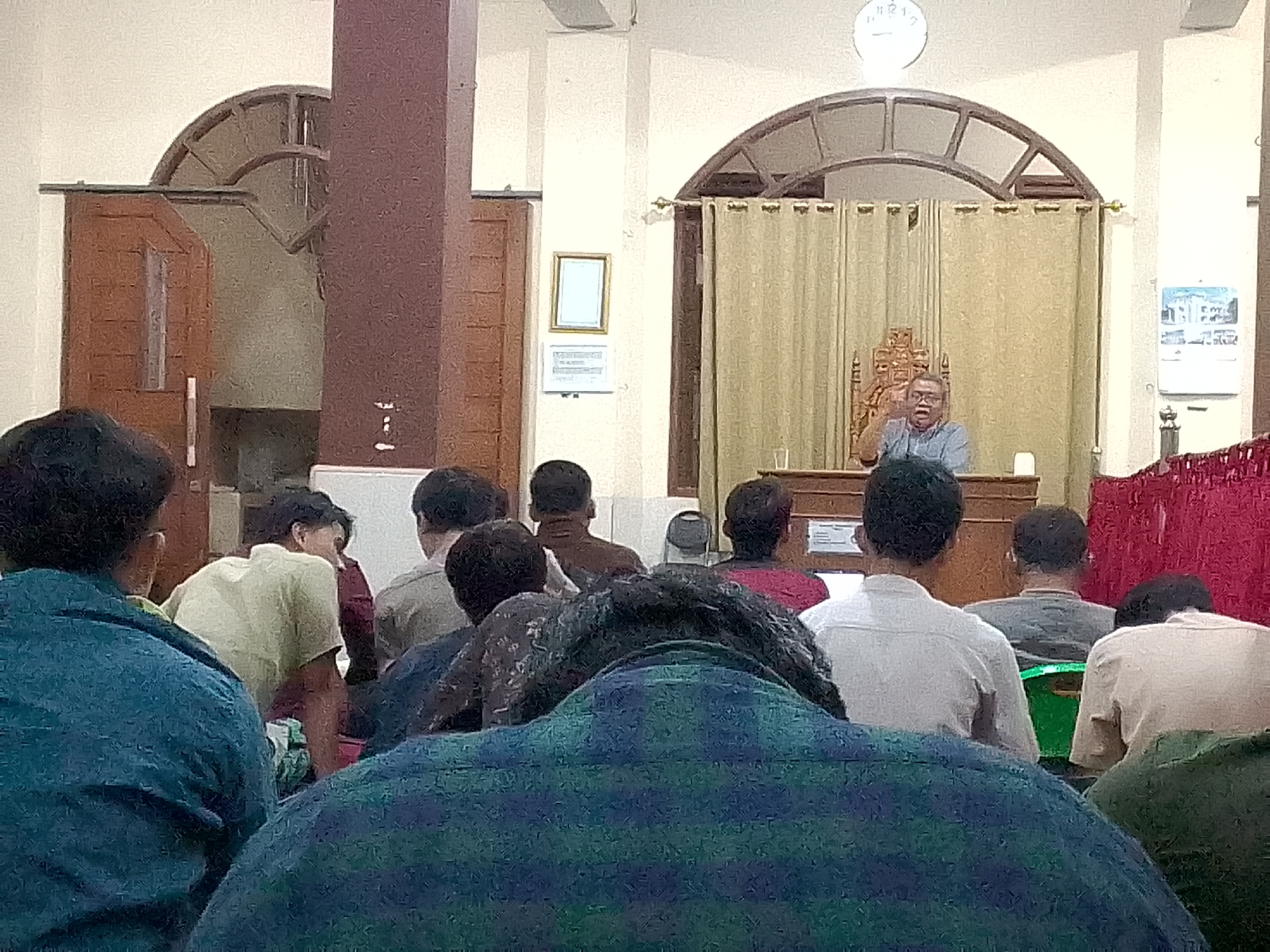 In the city of Kertajaya, Indonesia, a group of people are gathered in a room that appears to be a place of worship or a community hall. In the background, a man wearing glasses and a blue shirt is speaking from behind a wooden lectern, gesturing with his hands. He is seated on an ornate wooden chair. The room has a light-colored ceiling with a large arched window structure at the top. Beige curtains hang behind the speaker. On the left side of the room, there are wooden doors and a dark brown pillar. On the wall, a framed picture and a certificate or document are visible. The foreground shows the backs of several individuals, facing away from the camera, seated in rows. Their clothing consists of various colors, including blue, green, beige, and gray. The lighting in the room is ambient.