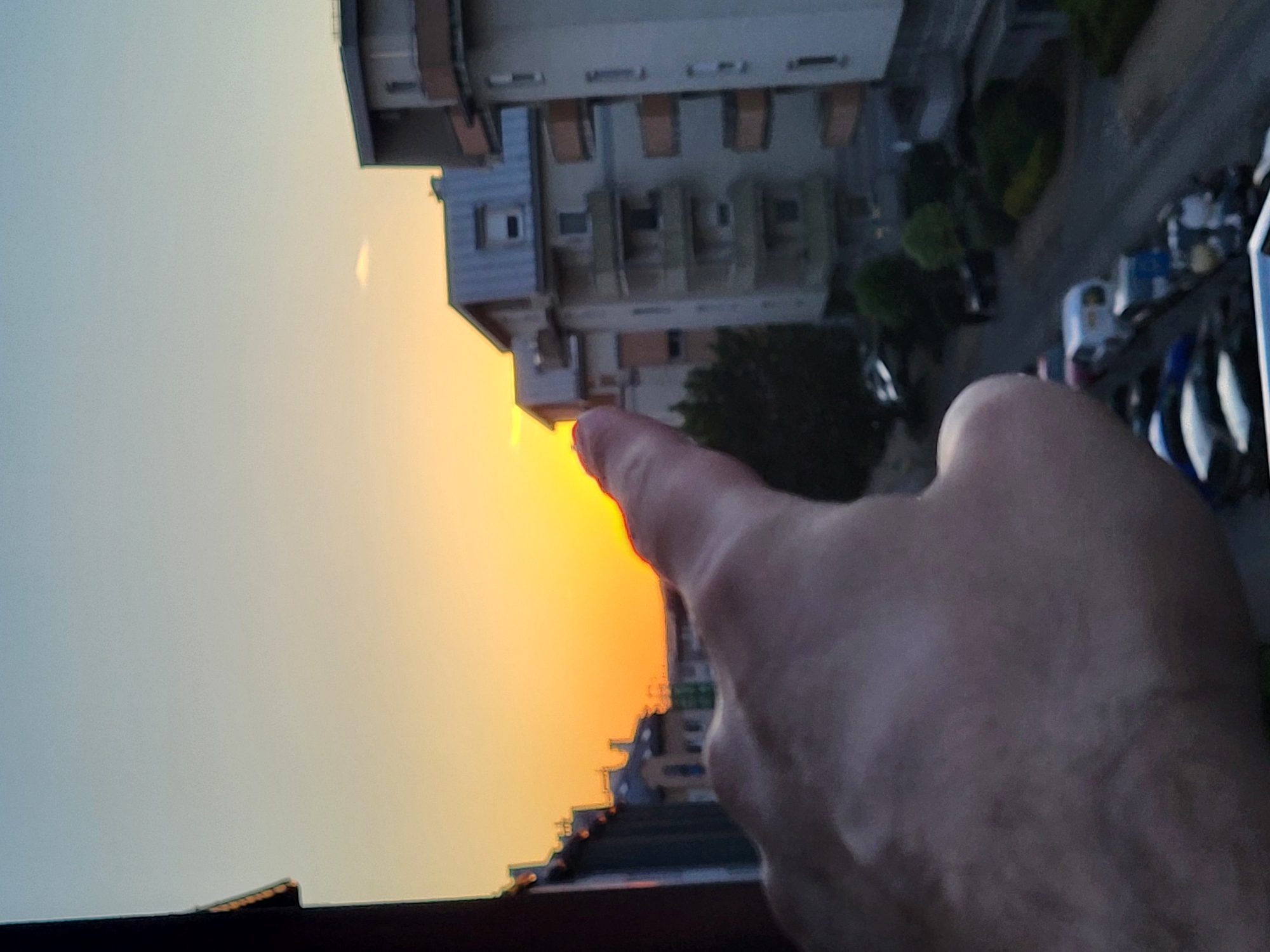 A close-up, low-angle photograph depicts a cityscape at sunset, with a hand in the foreground obscuring part of the view. The sky transitions from light blue to orange near the horizon. Several multi-story buildings are visible, with architectural features such as balconies and windows. The hand is fair-skinned, with a finger pointed towards the sunset. In the upper right corner, parked cars and trees are visible. The location appears to be a residential area within a city. The city and country are unknown.