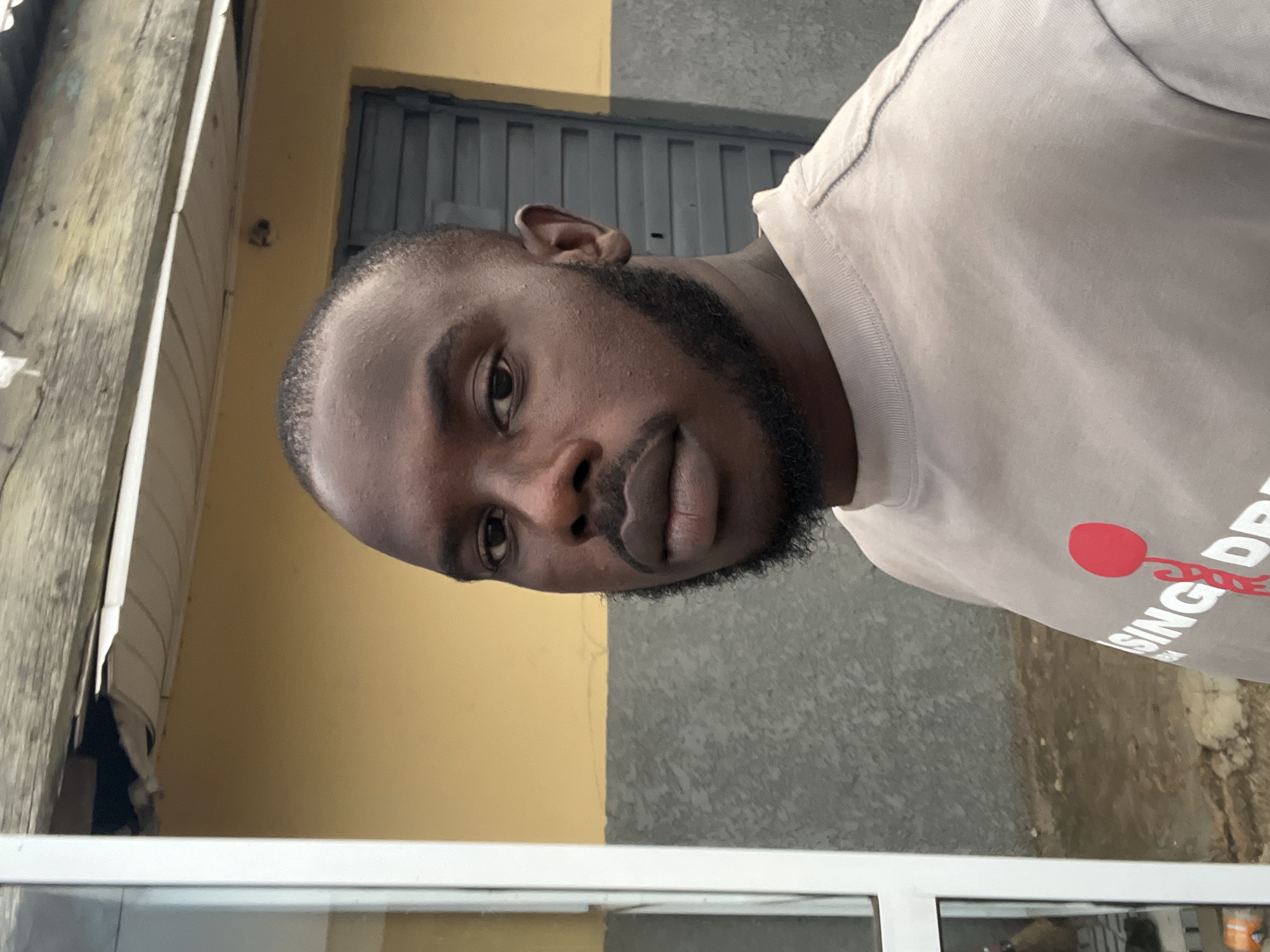 A close-up, angled portrait captures a man with dark skin, a short, neat beard, and short-cropped hair, looking directly into the camera. He wears a light-colored (possibly beige or pale grey) t-shirt or sweatshirt. A red circular graphic with an attached figure is visible on the shirt's right side, followed by white text that partially reads "SING DRI...". The man's expression is neutral and calm.
The setting appears to be an outdoor, urban environment in Jos, Nigeria, under bright, even daylight. The background is composed of various textures and structures. To the upper left, a weathered wooden structure or wall is visible. Directly behind the man's head, there is a section of a yellow-painted wall with a dark grey corrugated metal gate or shutter. Further to the right, a textured grey concrete wall serves as another part of the backdrop. The bottom of the frame shows a white horizontal element, possibly a window frame or a counter, reflecting light.