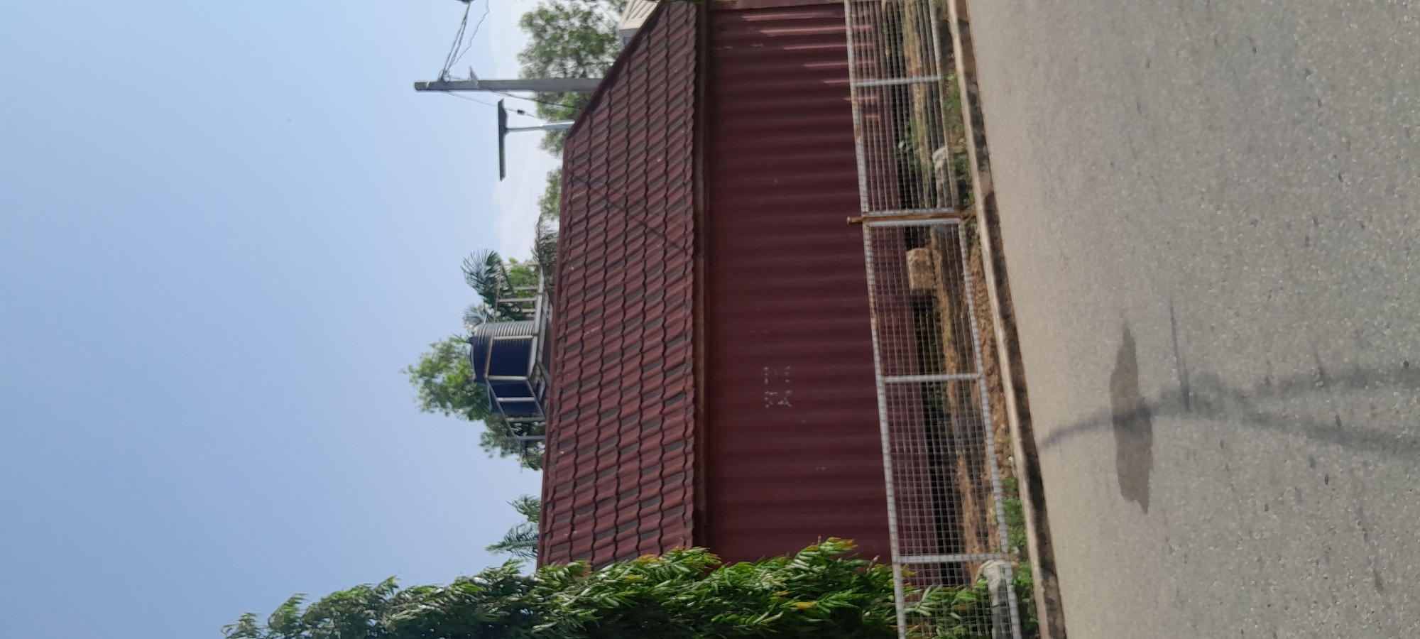 The image shows a view of a red corrugated metal building with a red tile roof, located on a street in Jalingo, Nigeria. A dark water tank is visible on top of the building. A metal chain-link fence runs along the front of the property, separating it from the road. A shadow of a utility pole stretches across the paved road in the foreground. Green trees are present to the left of the building and behind it. The sky is clear and blue.