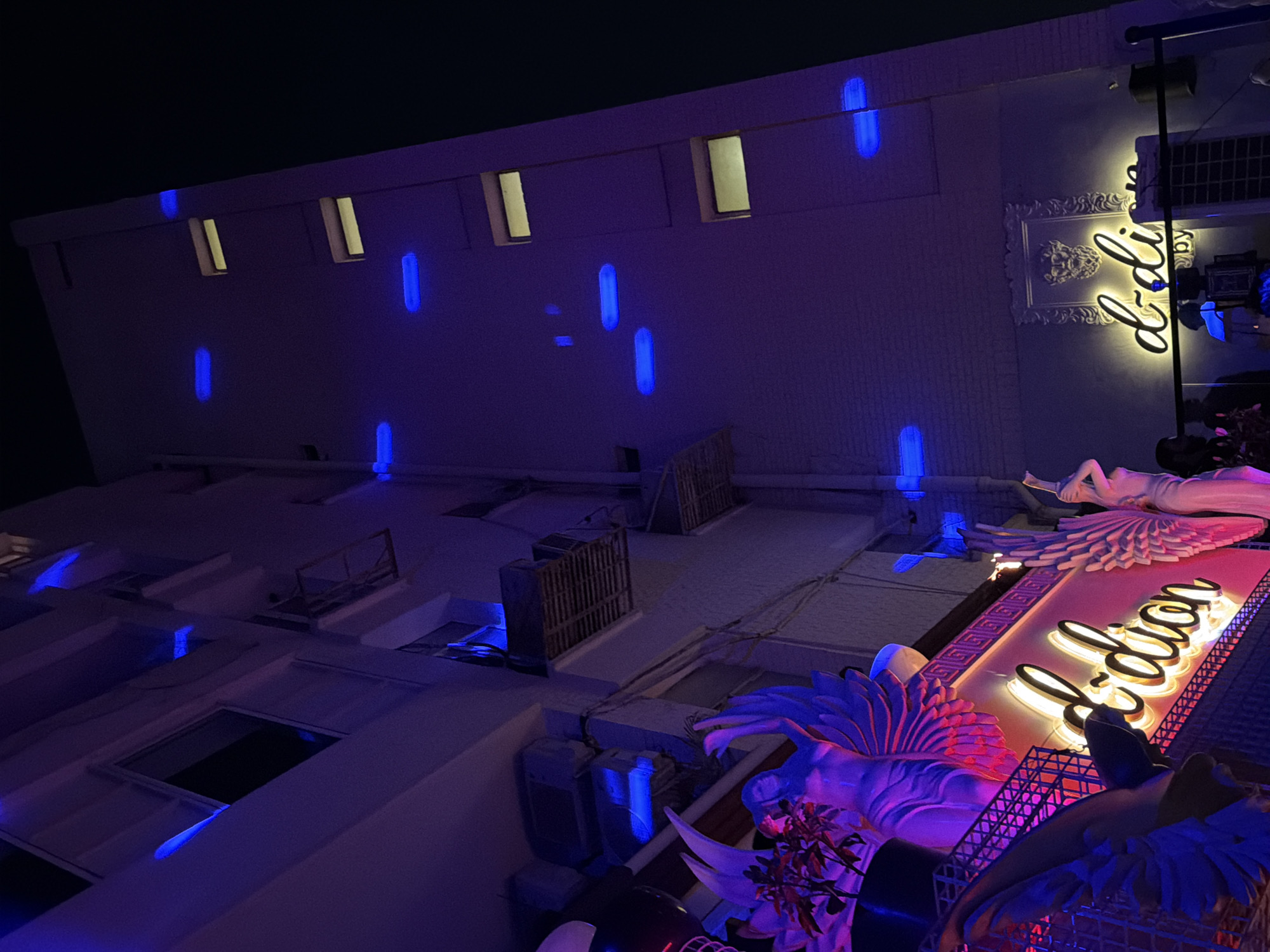 This image depicts the facade of D'Dion, a bar and lounge located in Safdarjung Enclave Market, South Delhi, India, observed at night.
The multi-story building, constructed of light-colored brick or concrete, is dramatically illuminated with artificial light, rendering its surface predominantly purple and blue. The upper sections of the facade feature numerous horizontally oriented blue neon or LED light strips, irregularly spaced. Multiple windows are visible; some are dark, while others exhibit a faint yellow internal light. Exterior air conditioning units are mounted on the building's left side.
The lower portion of the building is bathed in vibrant pink and magenta light. On the lower left, a prominent balcony or ledge displays at least two large, ornate statues of winged figures, illuminated in purple and pink. A plant with red leaves is situated near one of these statues. Above this area, a geometric frieze pattern is visible, also illuminated in pink.
Two main signs are present: a large, yellow neon script sign on the left reads "d-dion" against a pink background. A smaller, similar yellow neon script sign on the right reads "d-dion by". Above the smaller sign, an ornate, framed relief sculpture of a classical-style head or bust is mounted, illuminated by the ambient purple light. Black metal railings define the lower boundary of the visible area. Indistinct silhouettes of a few individuals are faintly discernible behind these railings on the lower level, predominantly on the right.