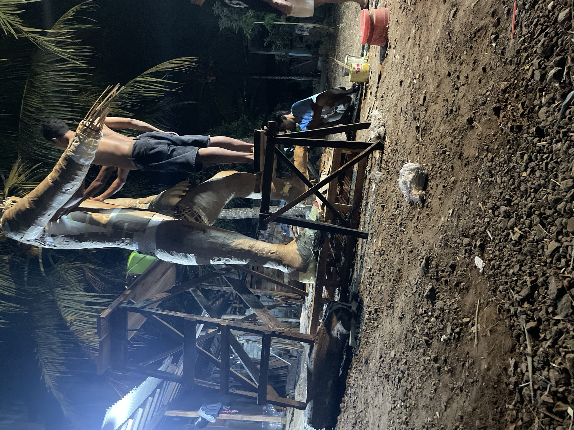 The image depicts an outdoor scene at night, likely in Kertosari, Indonesia, where a large effigy or sculpture is being constructed. In the foreground, the ground is covered with dirt and gravel. A massive, unfinished effigy, appearing to be human-shaped with a large leg and torso, stands center-right, supported by wooden scaffolding. Two individuals are actively working on the effigy. One, shirtless and wearing dark shorts, is positioned on a platform, appearing to be attaching thin wooden sticks to the effigy's outstretched arm. Another individual, wearing a blue shirt, is seated on the ground to the right of the effigy, observing or resting. Further right, a red bucket and a yellow paint can are visible on the ground. In the background, palm fronds hang from trees, and partially constructed wooden structures and what appears to be corrugated metal roofing are present. The lighting suggests artificial illumination, casting long shadows and highlighting the construction process.