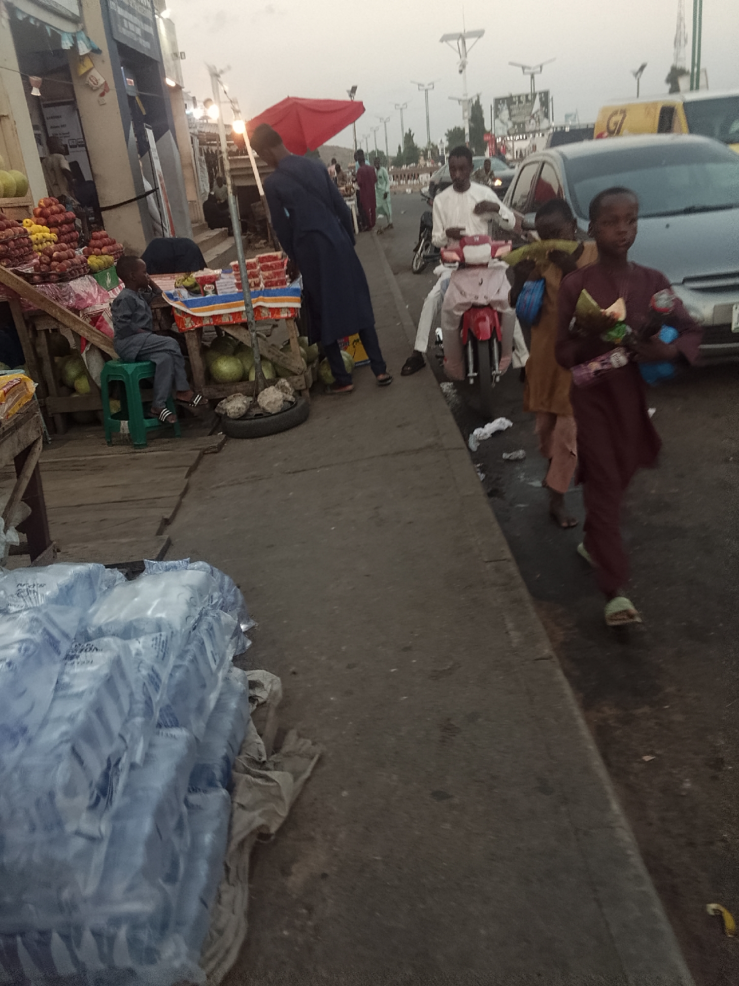 The image depicts a street scene in Pantame, Nigeria. A stack of water bottles wrapped in plastic sits on the left edge of the frame, near the sidewalk. Further down the sidewalk are vendors selling fruits and other goods under a red umbrella. One vendor is standing, while another is seated on a green stool. On the right side of the image, a road is visible. A man on a moped is present on the road along with two young boys in brown robes walking along the side of the road. In the background, other people, vehicles, and street lights can be seen.
