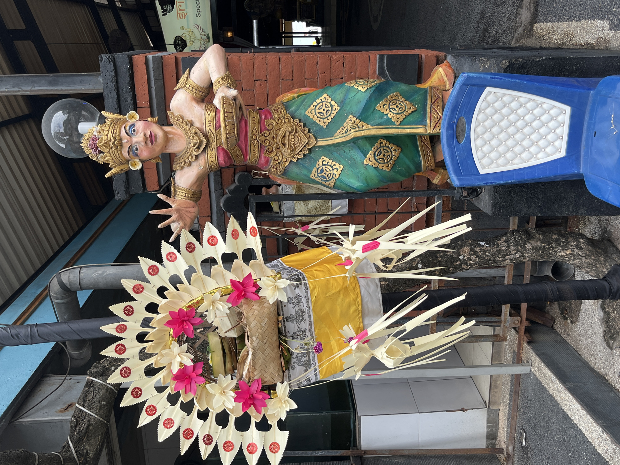 An exterior shot captures a colorful statue of a Balinese dancer adorned with a golden headdress and jewelry, wearing a pink top and a green skirt with gold embellishments. The statue stands in front of a brick pillar. To the left of the statue, a traditional Balinese offering, known as a canang sari, is displayed on a tall, ornate stand decorated with white and pink flowers and woven palm leaves. The offering is wrapped in a yellow and white cloth. A blue plastic chair is placed in front of the statue. The background reveals a building with a tiled roof, a metal pipe, and a glimpse of a restaurant sign in an Indian script. The scene is set outdoors in the city of Kuta, Indonesia, with a pavement and tree trunk visible in the foreground.