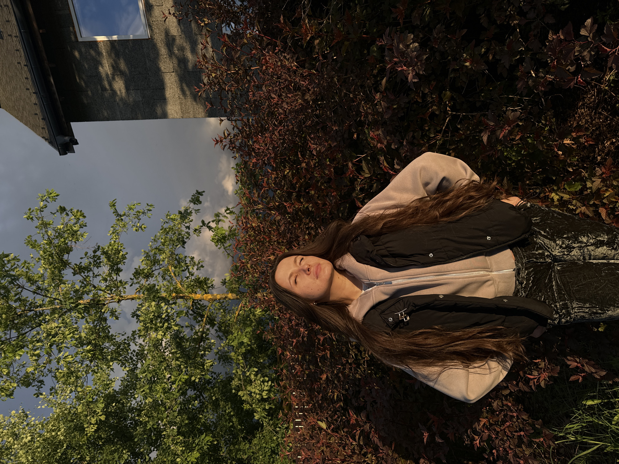 A young woman with long brown hair is lying amongst red and green foliage near a building. She is wearing a light-colored hooded sweatshirt with a black vest over it. The photograph appears to have been taken outdoors, possibly in a garden or yard. A section of a building with a window is visible, along with sections of trees and sky. The picture seems to be taken at sunset due to the lighting. There is no evidence within the image to confirm the location is Vysokinichi, Russia.