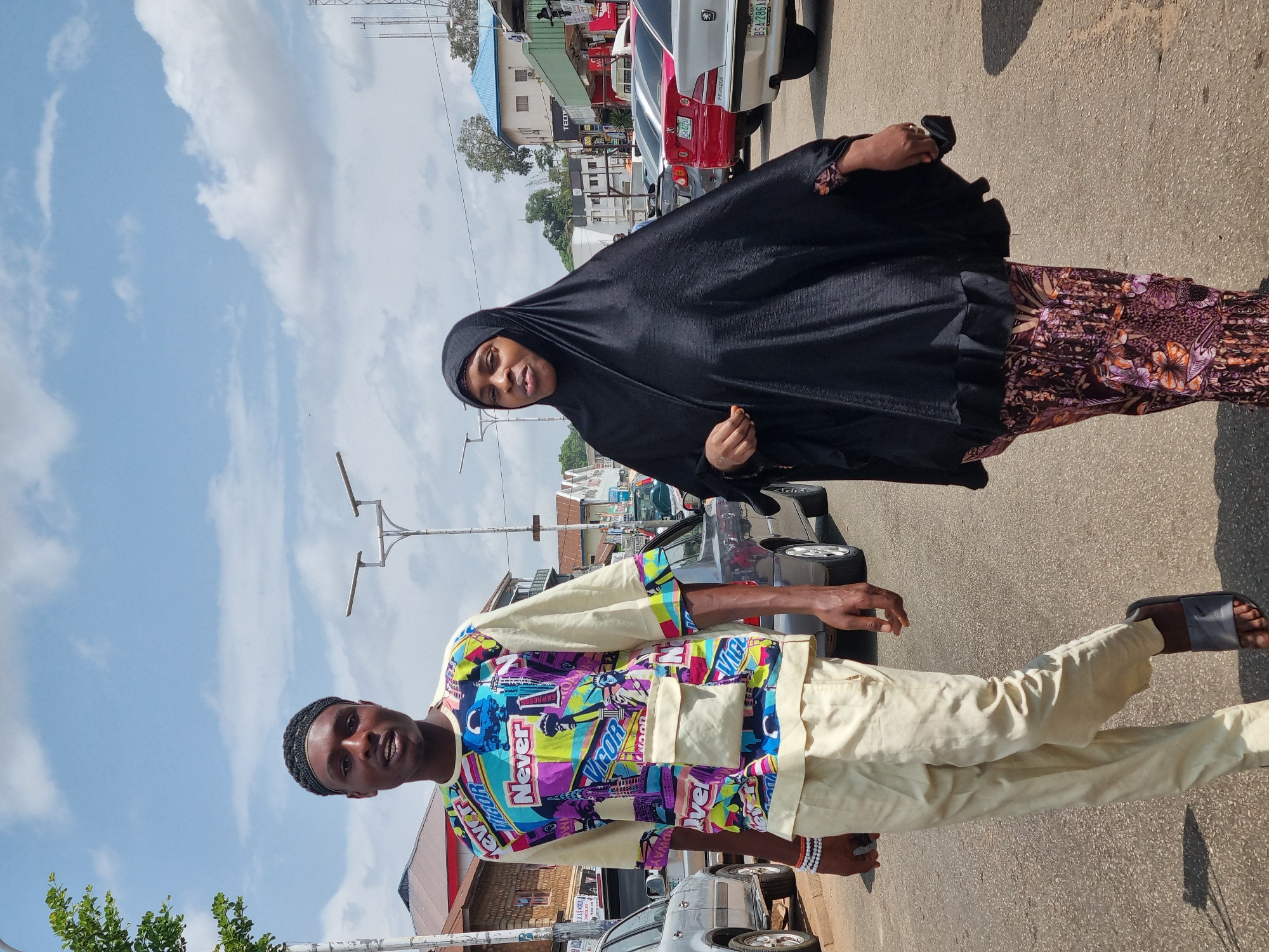 A man and woman are standing on a street in the city of Jos, Nigeria. The woman is wearing a black hijab and a floral dress. The man is wearing a colorful patterned shirt and light-colored pants. They are standing near parked cars on a street with buildings in the background. The sky is cloudy.
