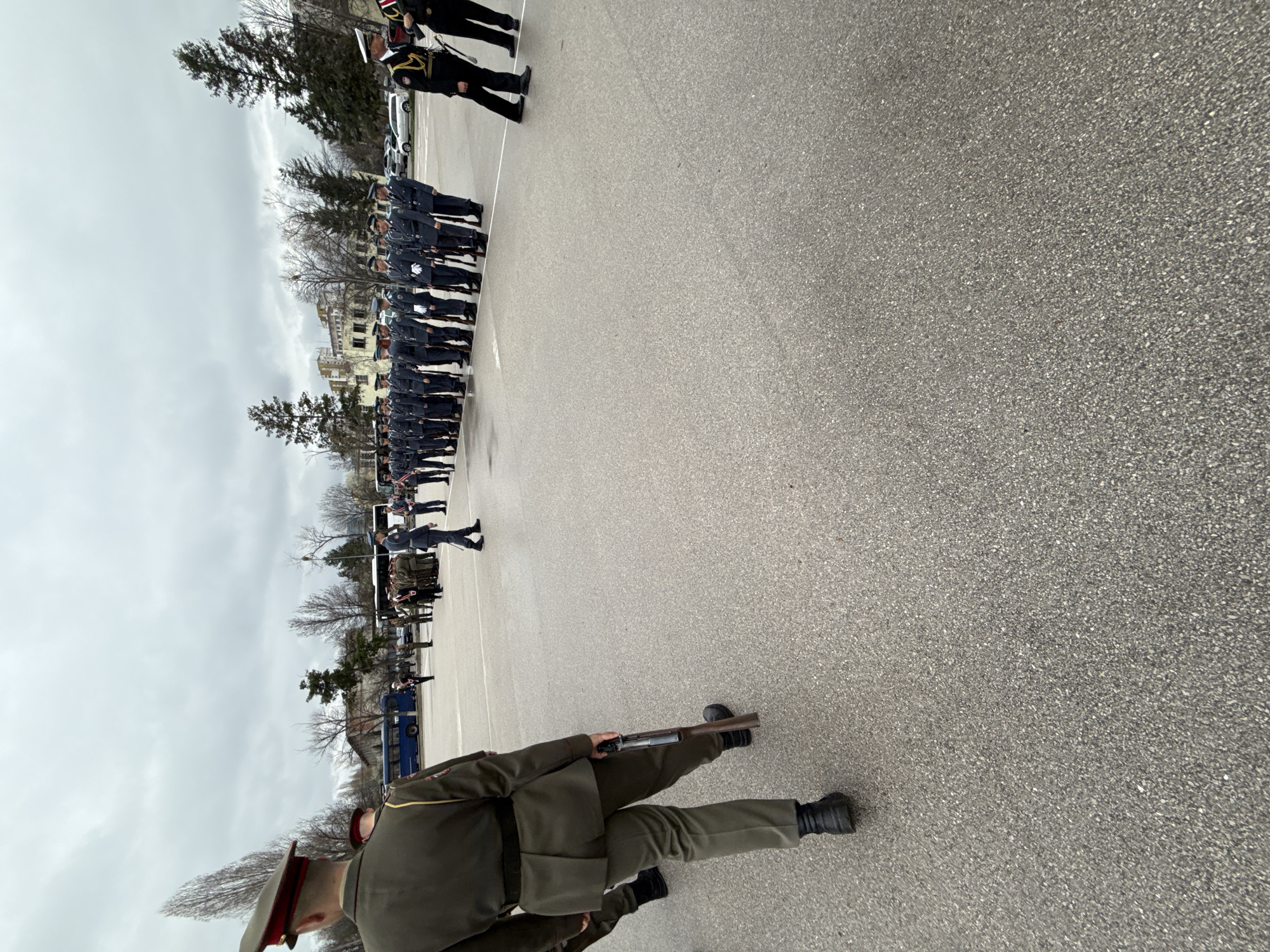 The image depicts a military parade or ceremony. A line of uniformed personnel stands at attention in the distance. A soldier in the foreground is holding a disassembled rifle. The setting is a large, paved area, possibly a parade ground, with buildings and trees visible in the background. The sky is overcast. The location could be Sofia, Bulgaria.