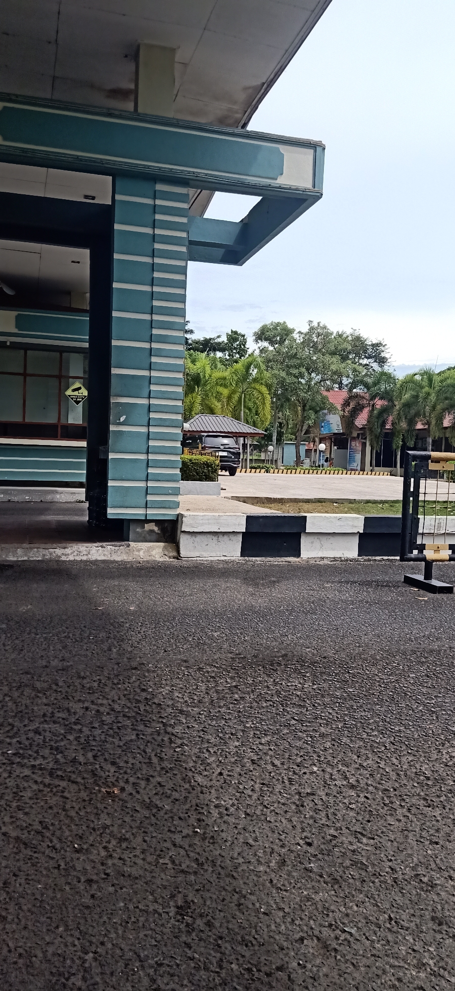 The image shows a paved road in front of a building with a distinctive teal and white striped facade. The building appears to be an office or government facility. In the background, there are palm trees and other vegetation, suggesting a tropical climate. A black SUV is parked under a small covered area. A black and white striped concrete barrier runs along the edge of the road. The sky is overcast. The overall scene depicts a functional, possibly administrative, area.