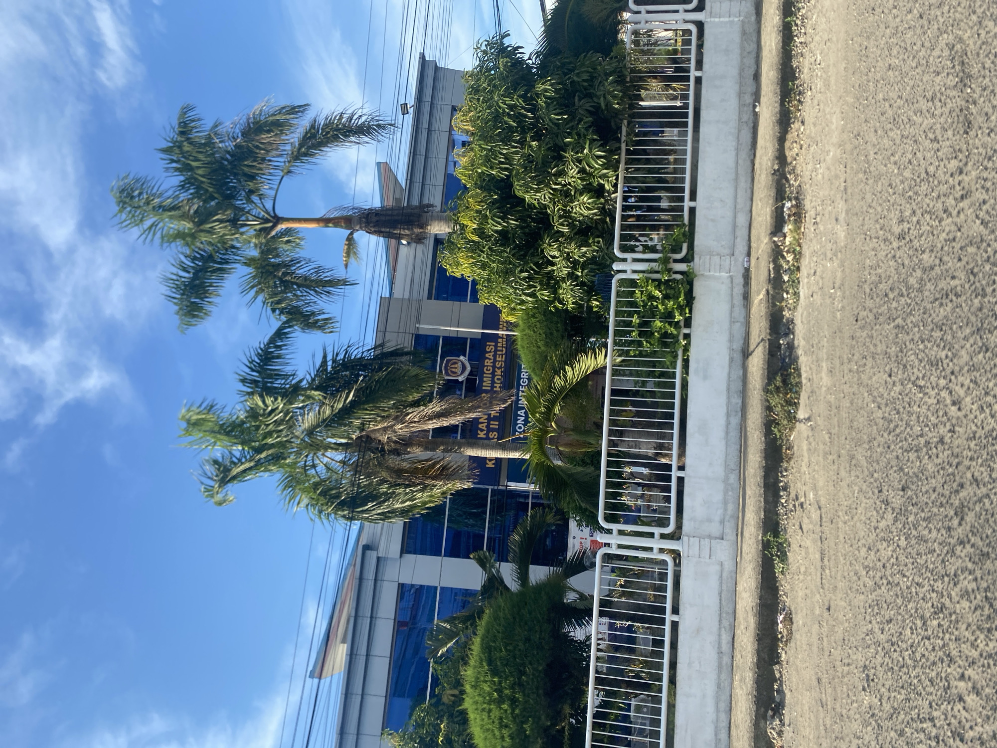 The image shows the Kantor Imigrasi Kelas II TPI Lhokseumawe (Lhokseumawe Immigration Office Class II) in Lhokseumawe, Indonesia. The building is modern, with blue glass windows and a sign with the office's name and logo. Two tall palm trees are in front of the building, and a white metal fence runs along the edge of the property, separating it from the road. The road is asphalt, and the sky is blue with scattered clouds.