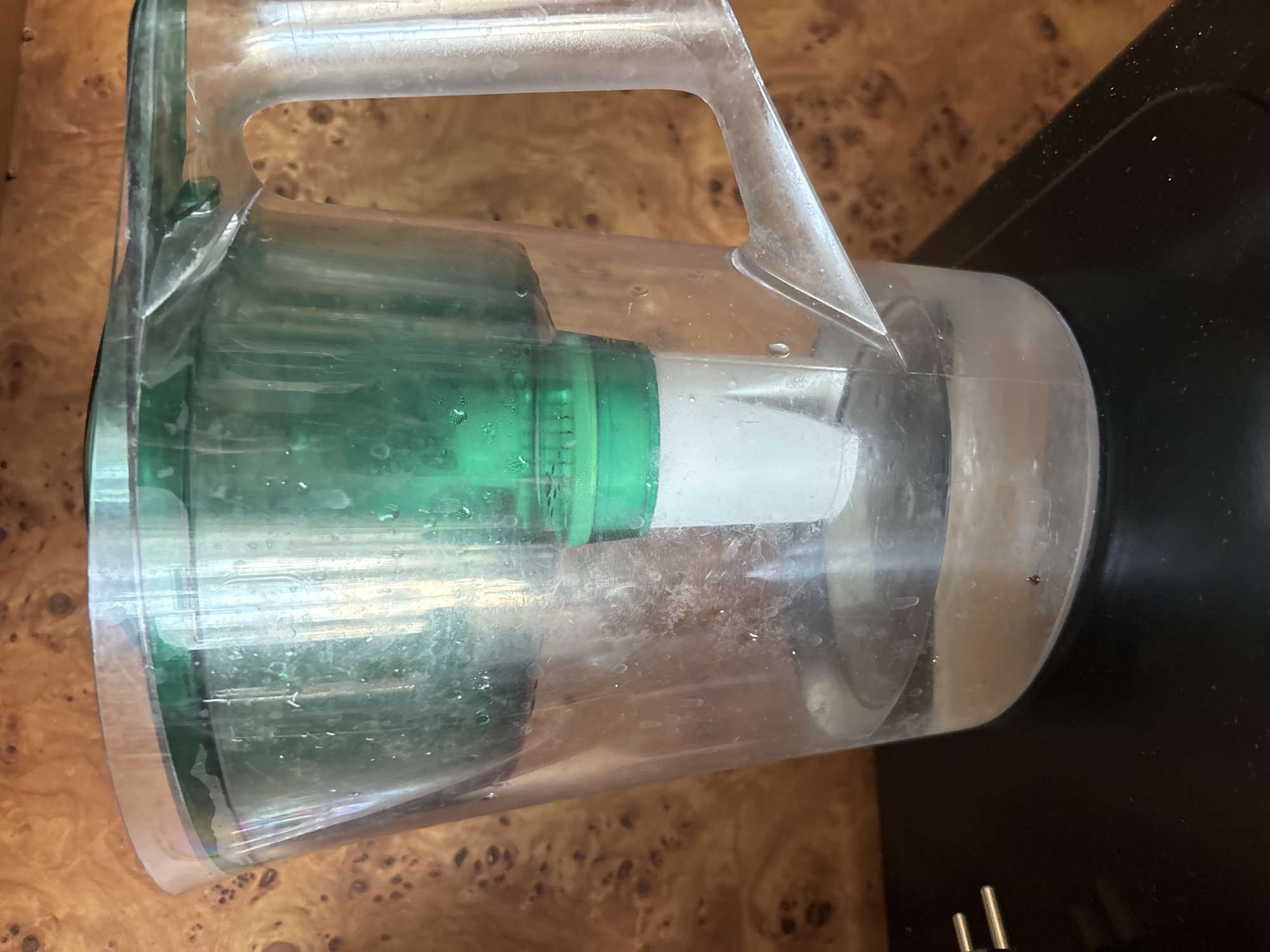 A clear plastic water pitcher with a green filtration system is positioned on a black surface, possibly a countertop. The pitcher contains water. The background is a wood-grain pattern, suggesting an indoor setting. The image was taken in Blagoveshchensk, Russia.