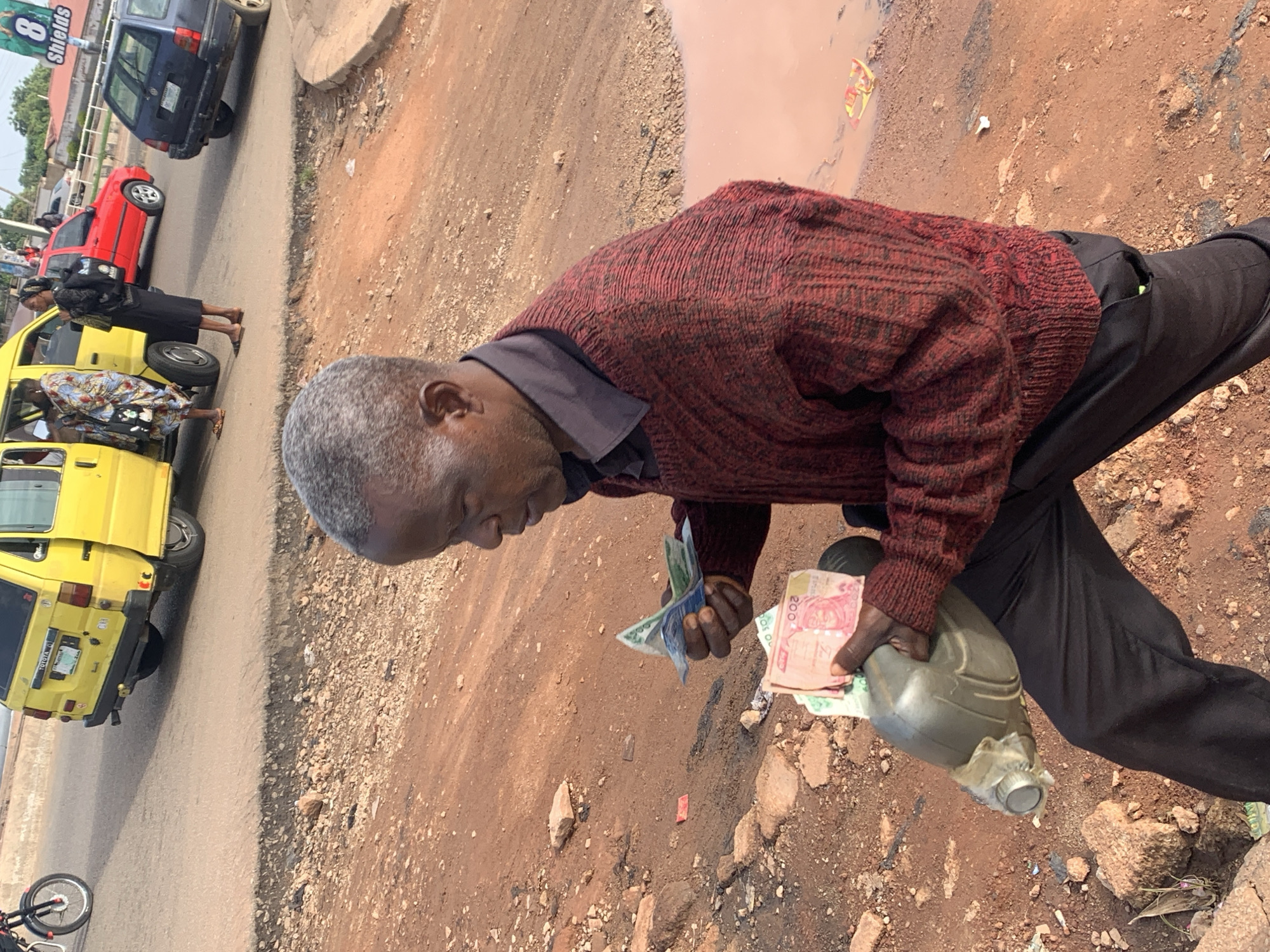 The image shows an outdoor scene in Gura Top, Nigeria. In the foreground, a man with gray hair is holding several banknotes, including a 200 naira note, in his right hand. He is wearing a dark maroon sweater with a textured pattern and black pants. He is also carrying a green plastic jerrycan in his left arm. The ground around him is made of reddish-brown dirt with scattered rocks and debris. In the middle ground, a yellow minivan with its rear door open is parked on a paved road. Two women are visible near the minivan. One woman, wearing a colorful patterned dress, is entering the minivan. Another woman, wearing a black skirt and headscarf, stands beside the open door. A red car and a dark blue car are also parked on the road behind the minivan. In the background, there are buildings and a billboard with the text "8 SHIELDS". The sky is partly visible and appears overcast. The overall scene depicts a busy street or roadside area with transportation and commerce.