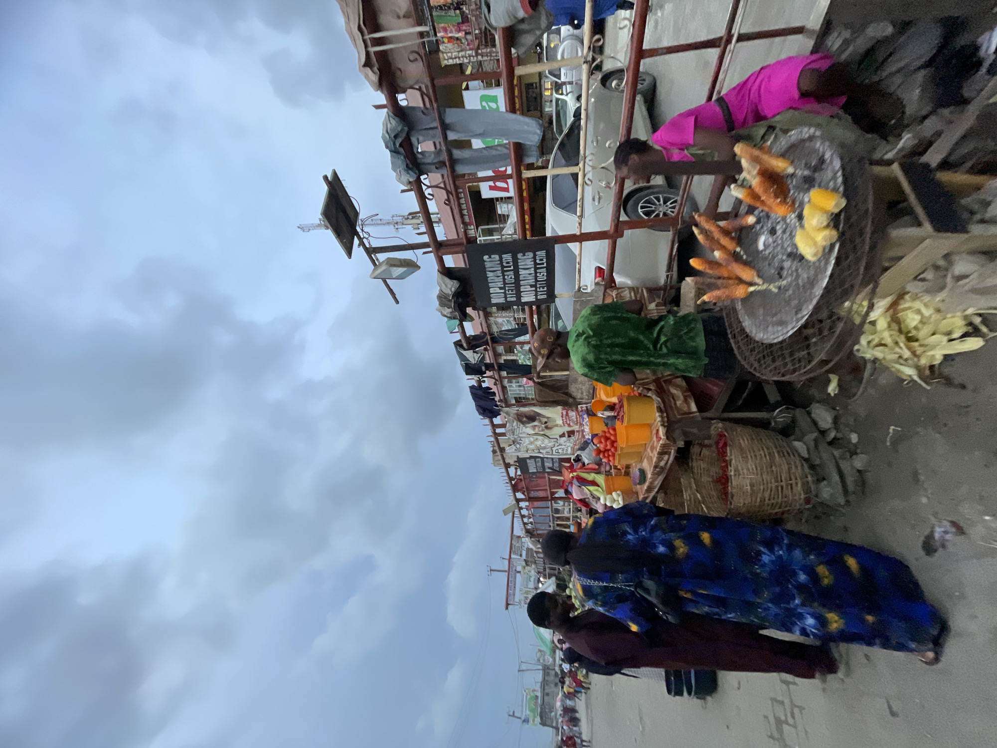 This image captures a bustling outdoor street market scene in Lekki, Nigeria, under an overcast sky. The photograph is taken from a slight upward angle, revealing a row of market stalls constructed with metal frames.
Numerous individuals are present, engaged in various activities typical of a market. A central focus is a person wearing a vibrant pink top, actively tending to several ears of corn roasting on a large, circular metal grill. This indicates the preparation and sale of street food. Adjacent to this, other stalls display fresh produce, including tomatoes and onions, arranged in plastic containers and traditional wicker baskets.
Other people are visible near the stalls, some appearing to be vendors and others possibly shoppers. In the foreground, the back of a person wearing a distinct blue patterned dress is seen. Behind the market stalls, vehicles, including a light-colored car, are parked or driving, suggesting the market runs alongside a road.
The sky is filled with grey, overcast clouds, indicating either a cloudy day or possibly the early evening. A prominent black sign attached to one of the market structures is clearly visible, bearing the text "NO PARKING" repeated, followed by "JETI OSA LCJM." Another partially visible sign appears to include the word "MARKET." The scene conveys a lively hub of local commerce and daily activity.