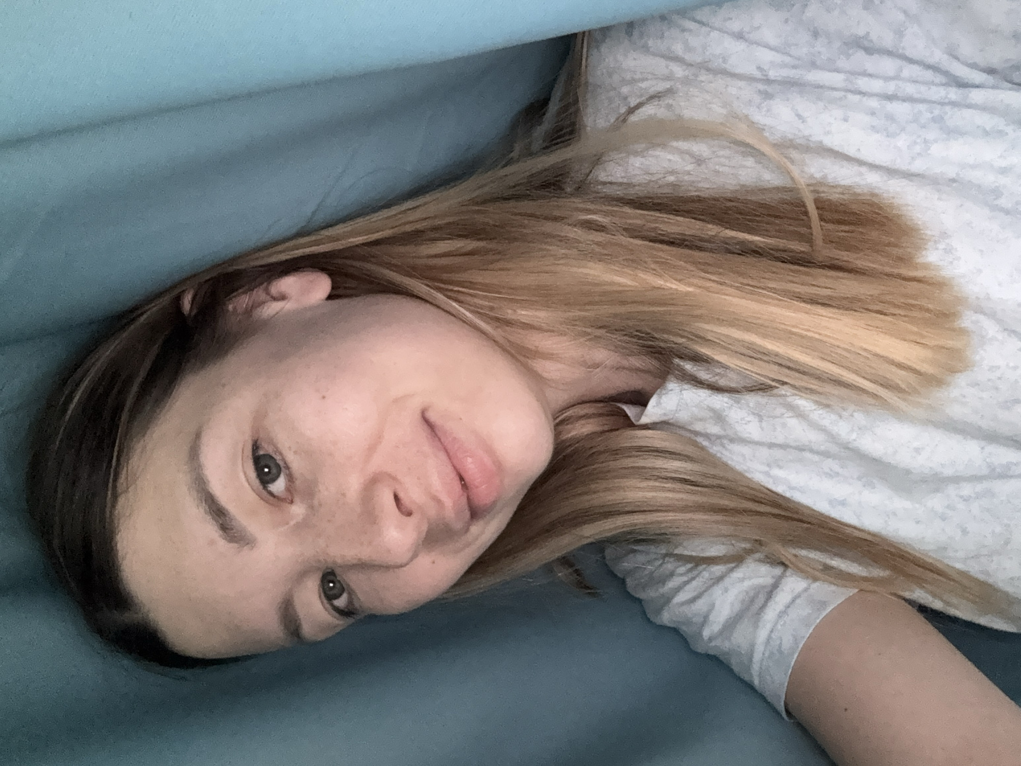 A young woman with light brown hair and fair skin, lightly freckled, is seen in a close-up, head-and-shoulders shot. She is wearing a light-colored t-shirt with a subtle pattern. The background consists of draped, teal-colored fabric. Her expression is neutral, with a slight smile. The image appears to be a selfie taken indoors. There is no identifiable information to place this image in Moscow, Russia.