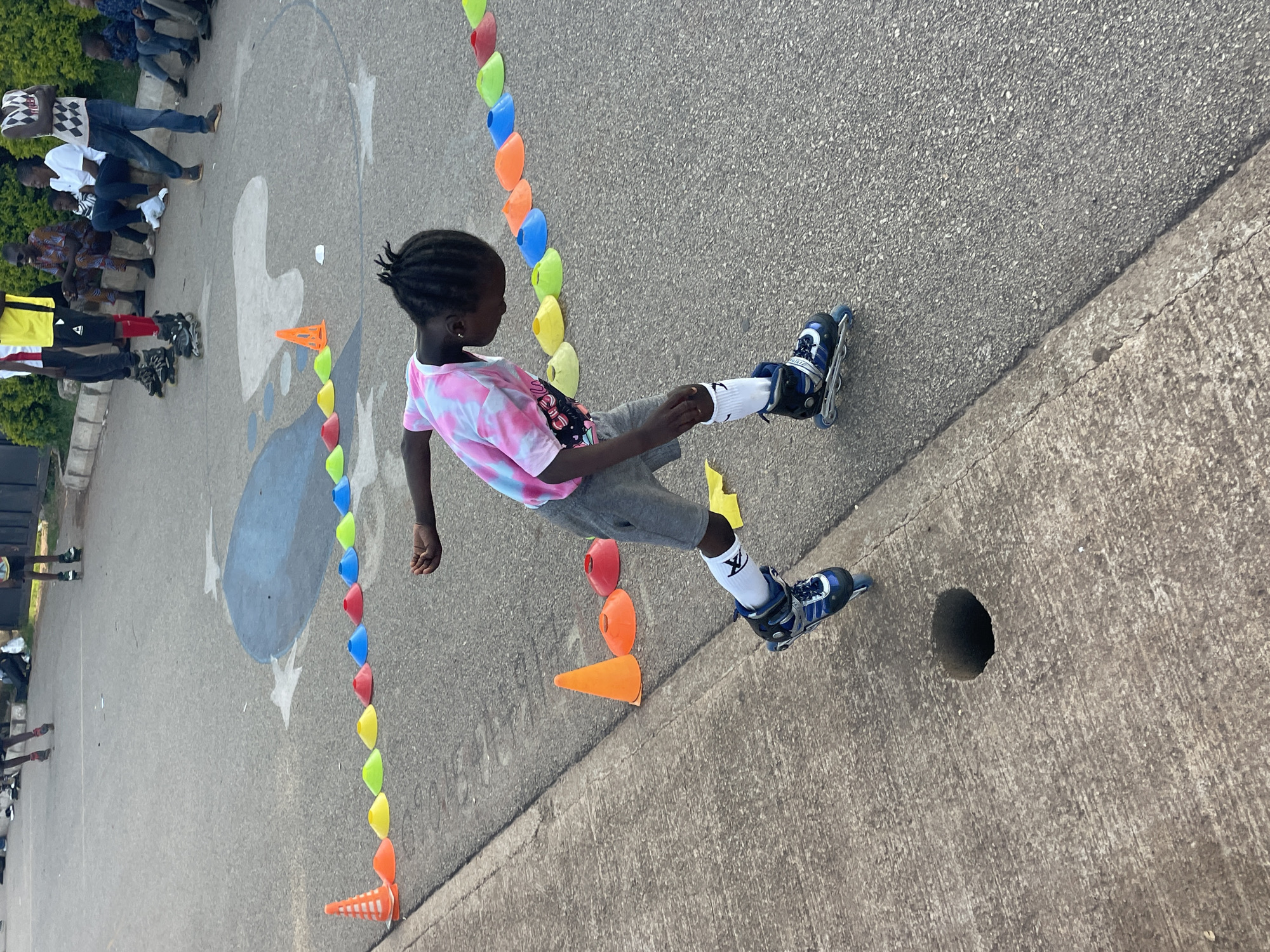 In Jos, Nigeria, a young Black child wearing rollerblades and white socks with black trim is engaged in a slalom exercise, maneuvering between a line of colorful cones set up on an asphalt surface. The child is dressed in grey shorts and a tie-dye t-shirt in shades of pink and white, with their dark hair styled in braids. The scene appears to be outdoors, with a concrete curb to the left of the frame, featuring a circular drain opening. In the background, other individuals are visible, some seated and others standing, observing the activity. The environment suggests a public space or recreational area, with a painted blue circular design on the asphalt in the mid-ground. The lighting indicates it is daytime.