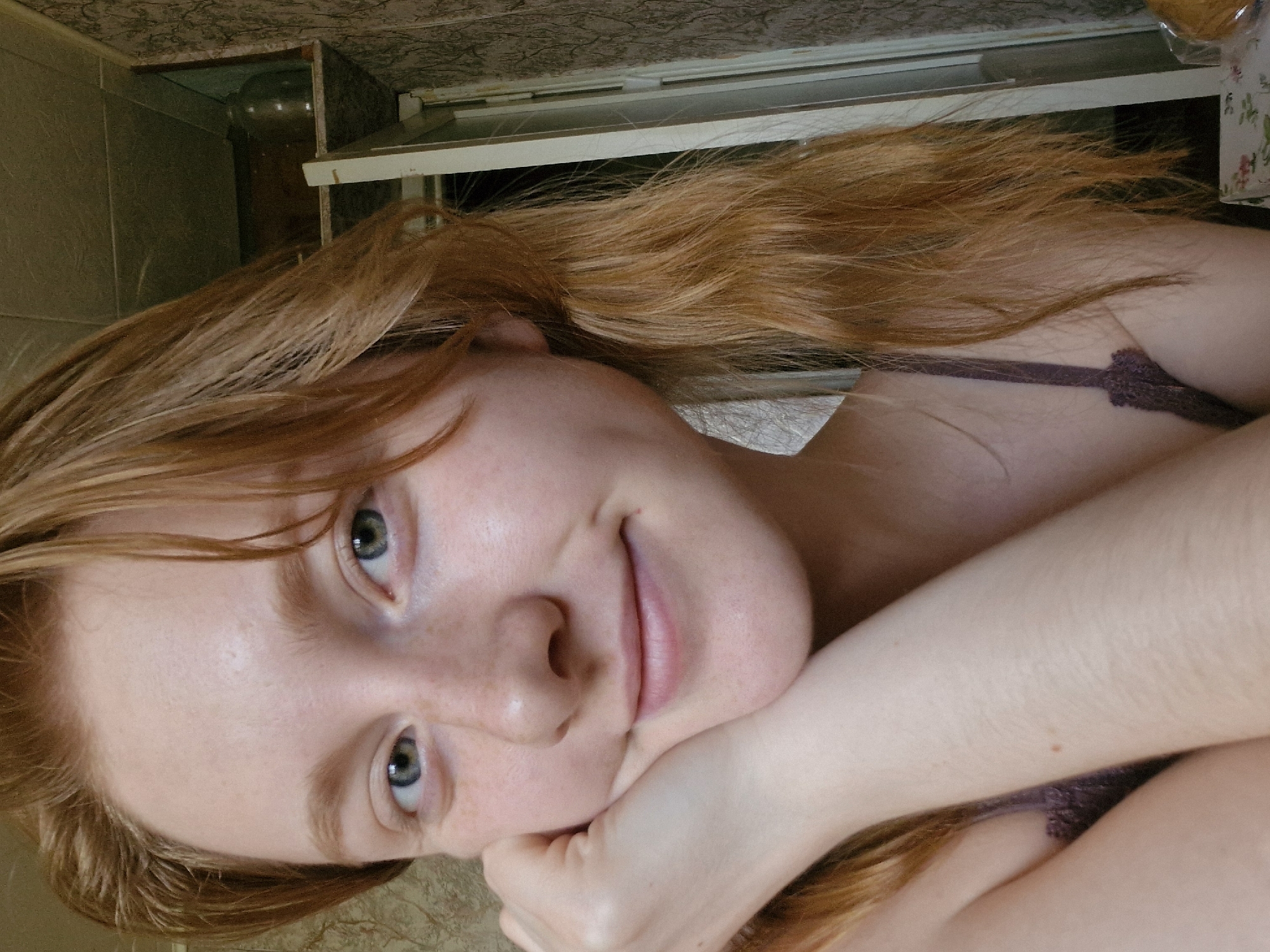 A close-up photograph depicts a young woman with light skin, reddish-blonde hair, and blue-green eyes. Her face is tilted towards the camera, with her hands resting beneath her chin. The woman has freckles visible on her face. She is wearing a dark-colored garment with lace detailing. The background includes tiles and a cabinet. The location in Yekaterinburg, Russia, is not discernible from the image.