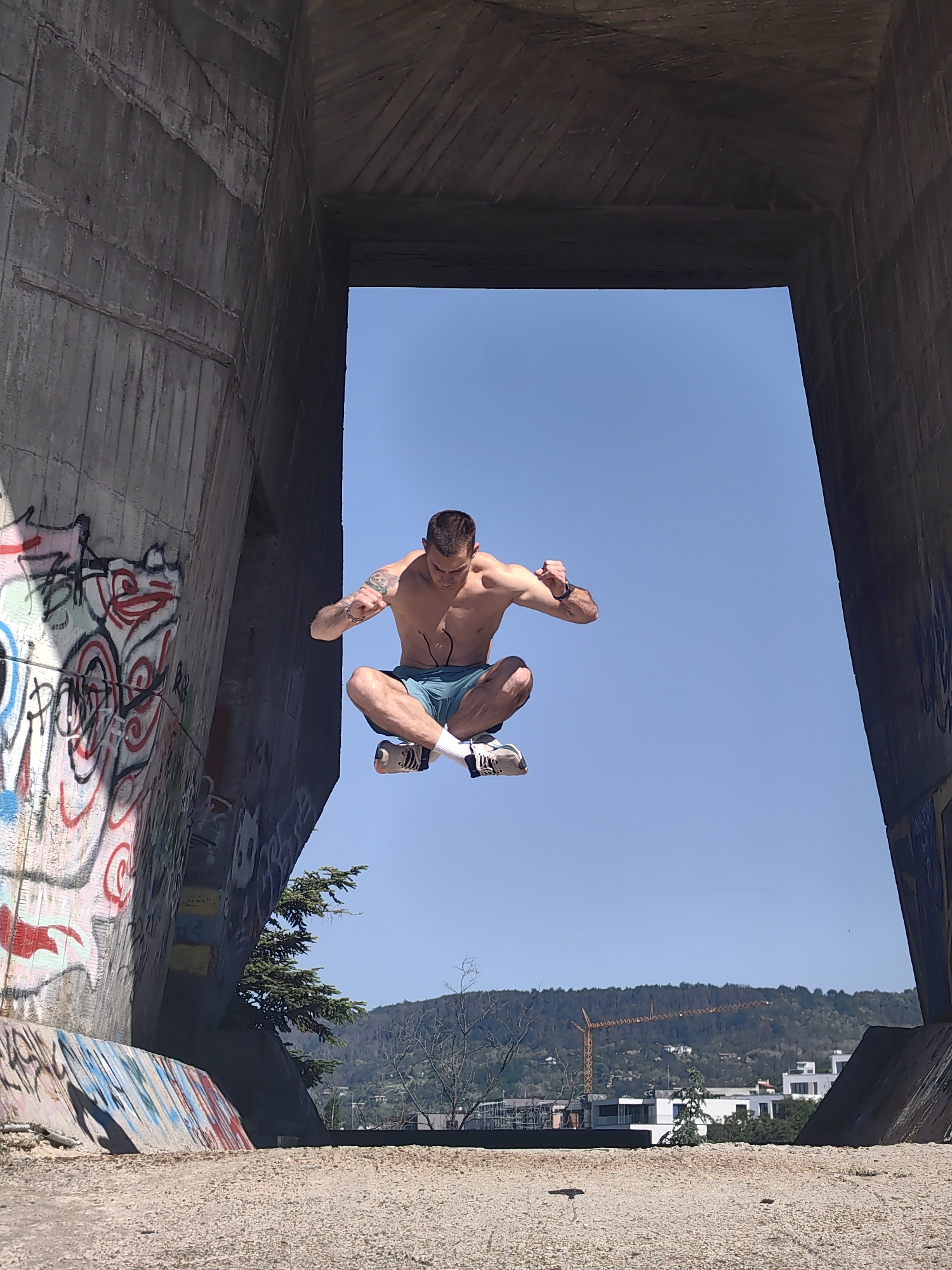 A shirtless man wearing blue shorts, white socks, and sneakers levitates in a lotus position within a concrete structure with graffiti on one side. The man has tattoos on his left arm and is clenching his fists. The structure frames a view of a blue sky, a hillside with trees, buildings, and a yellow crane. The photo was taken at Asparuhovo Bridge in Varna, Bulgaria.